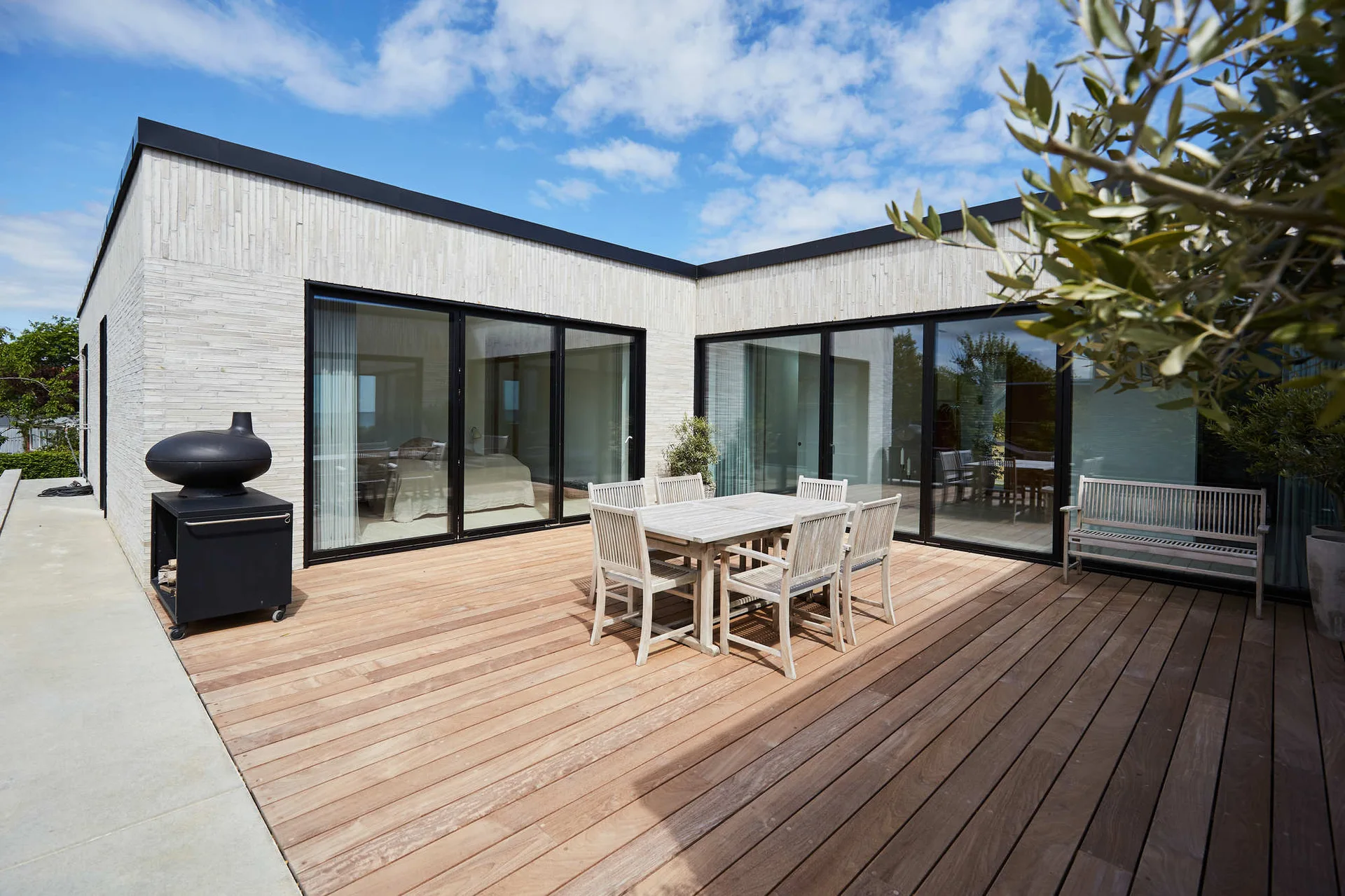 A modern outdoor terrace features a wooden dining table and chairs on a wood plank deck next to light brick walls and sliding glass doors.