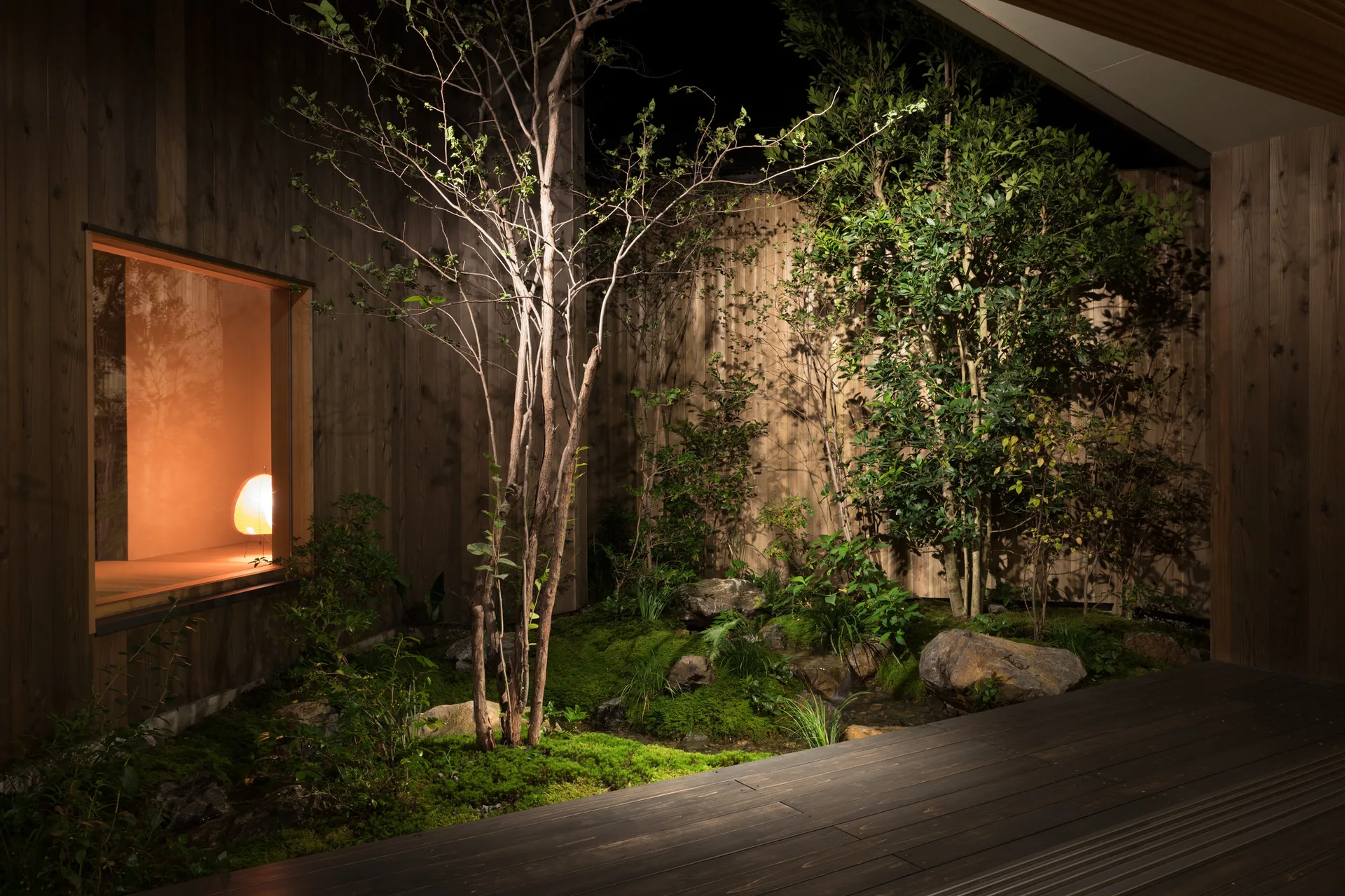 A dark wooden deck overlooks a small, illuminated courtyard garden at night with moss, rocks, trees, wood siding, and a glowing window.