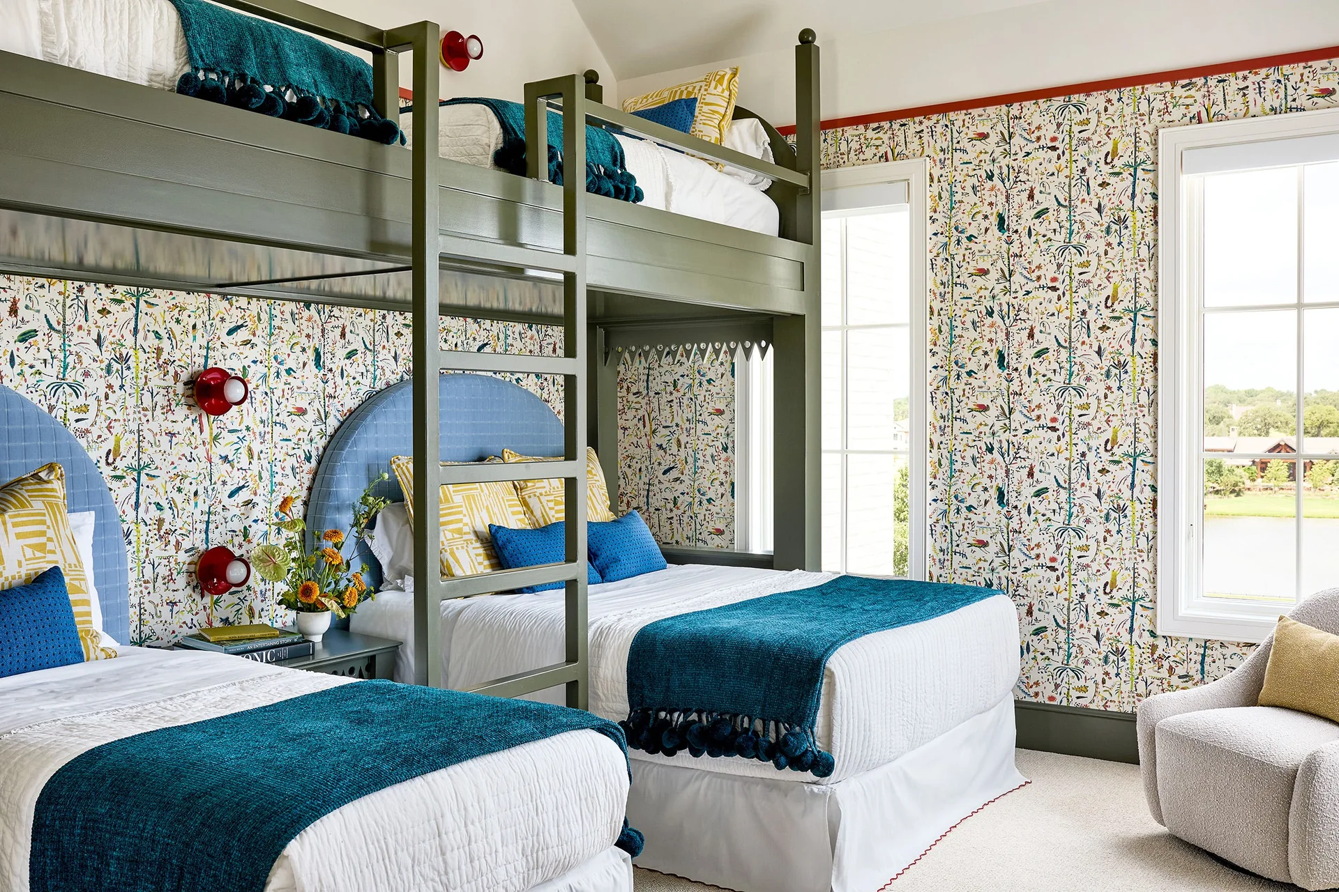 A kids bedroom features custom olive green bunk beds against botanical print wallpaper with blue headboards and teal throw blankets.