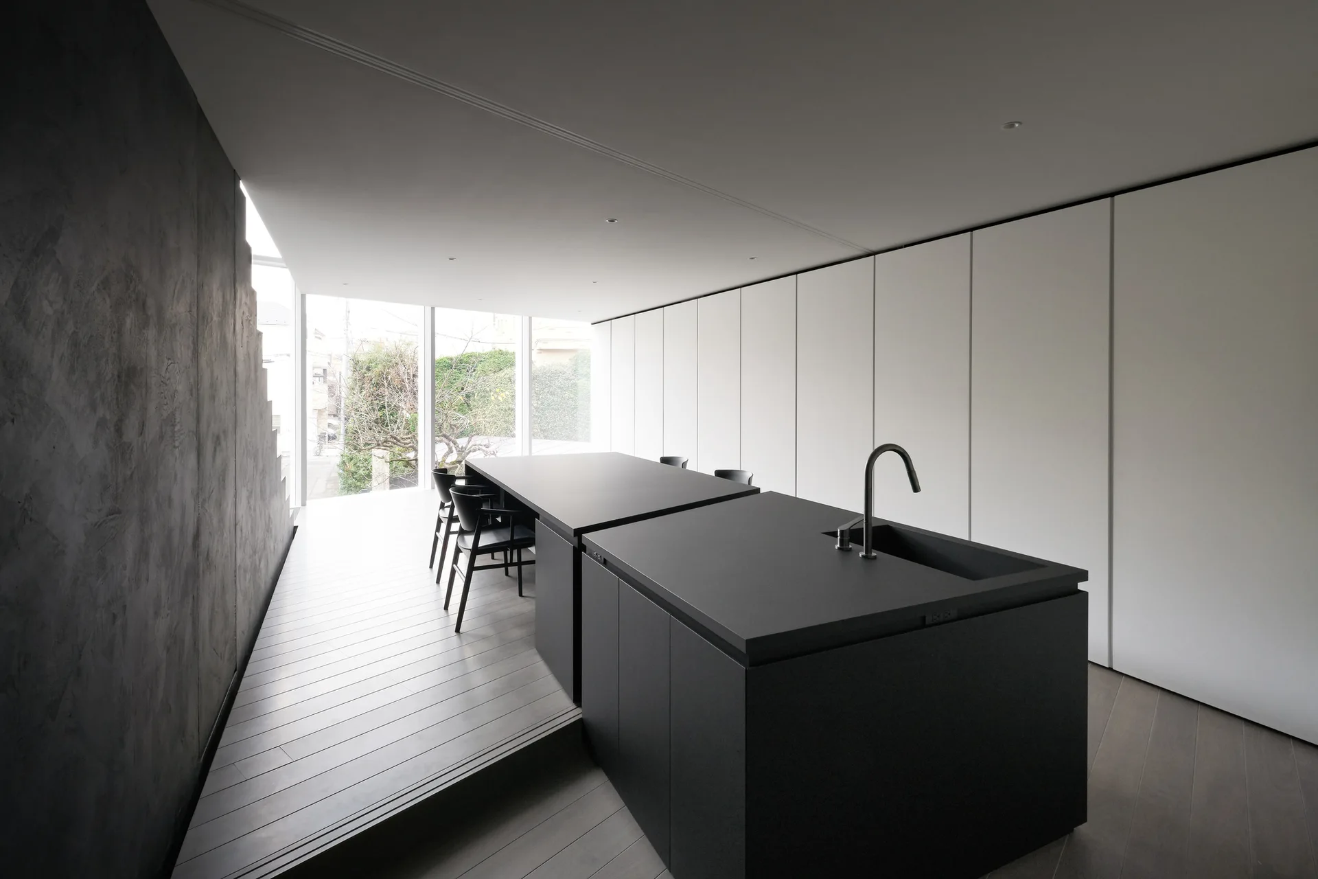 A minimalist kitchen features a matte black island, an integrated dining table, a textured grey wall, and a full-height white storage wall.