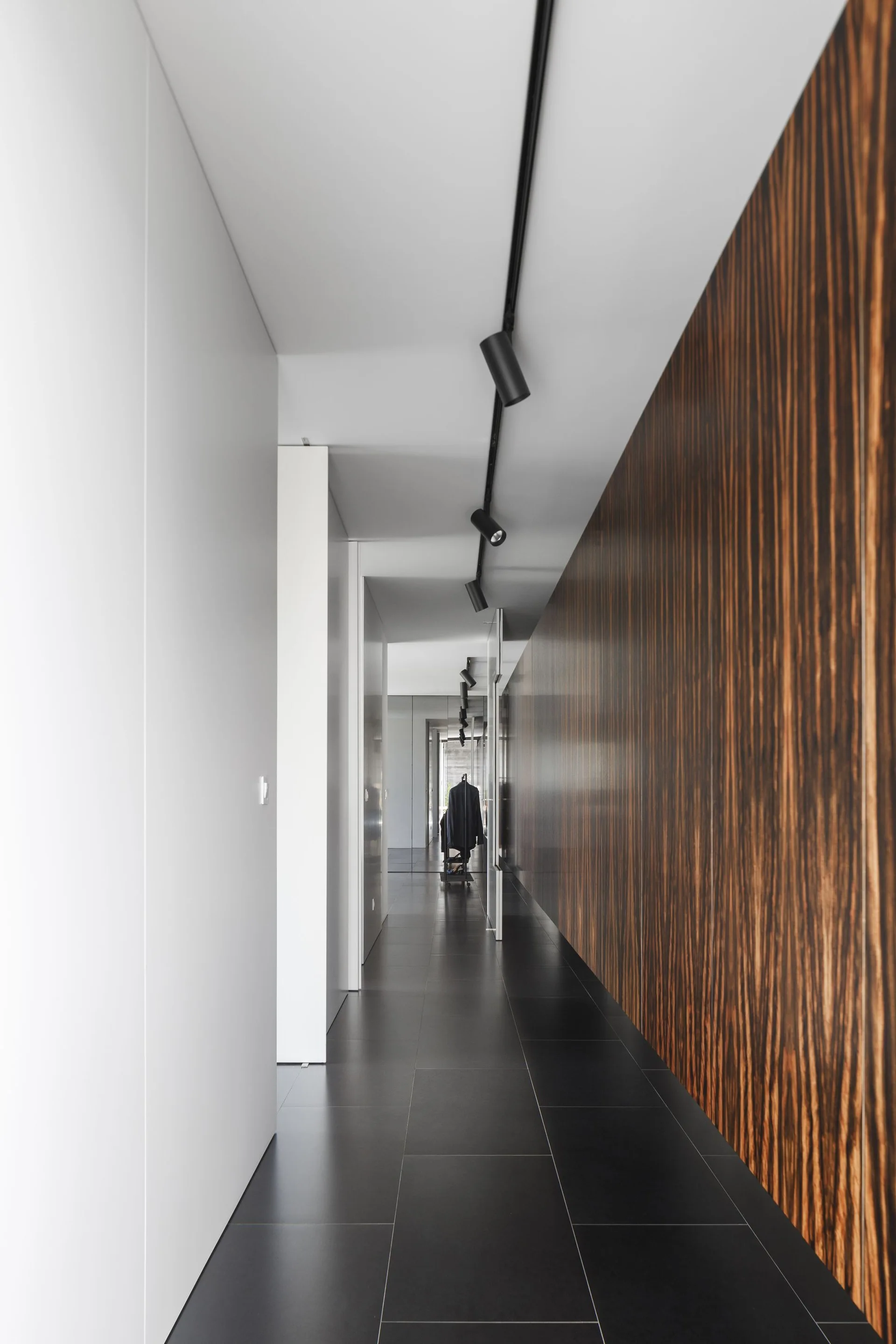 A long minimalist hallway features a dark vertical wood-grain paneled wall, dark floor tiles, white walls, and black ceiling track lighting.