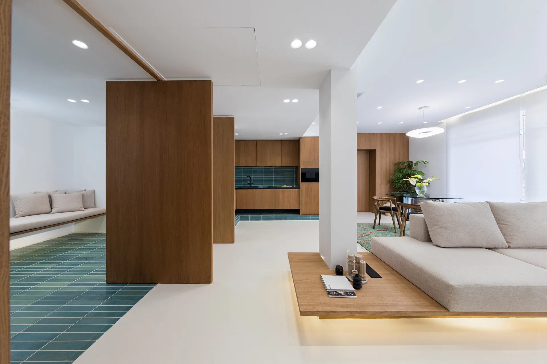 A modern open-plan living room featuring a low beige sectional sofa on a wood platform, teal tile flooring, and wood-paneled partition walls.