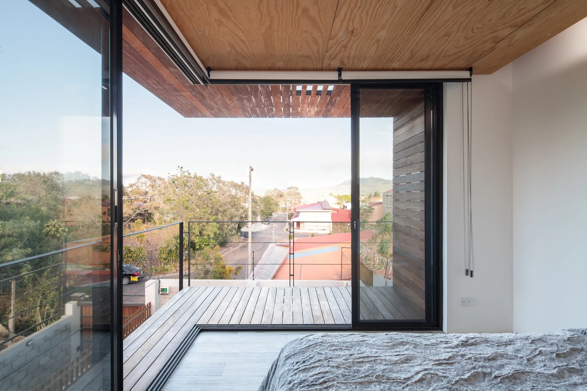 A bedroom with a grey textured bed features large black-framed sliding glass doors opening onto a wood-decked balcony with a thin metal railing.