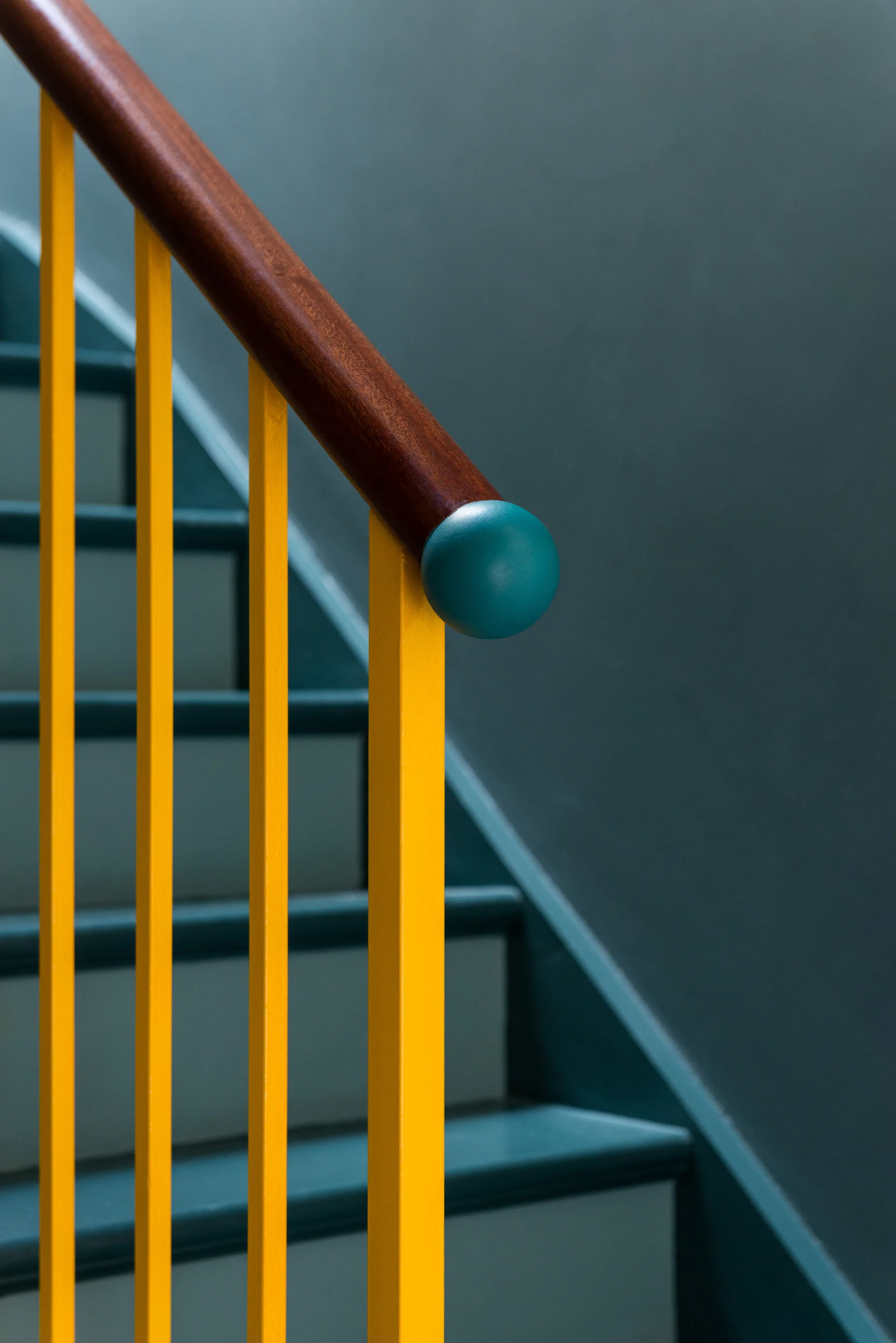 A close-up of a staircase showing bright yellow metal spindles, a polished wood handrail with a teal round finial, and dark teal steps.