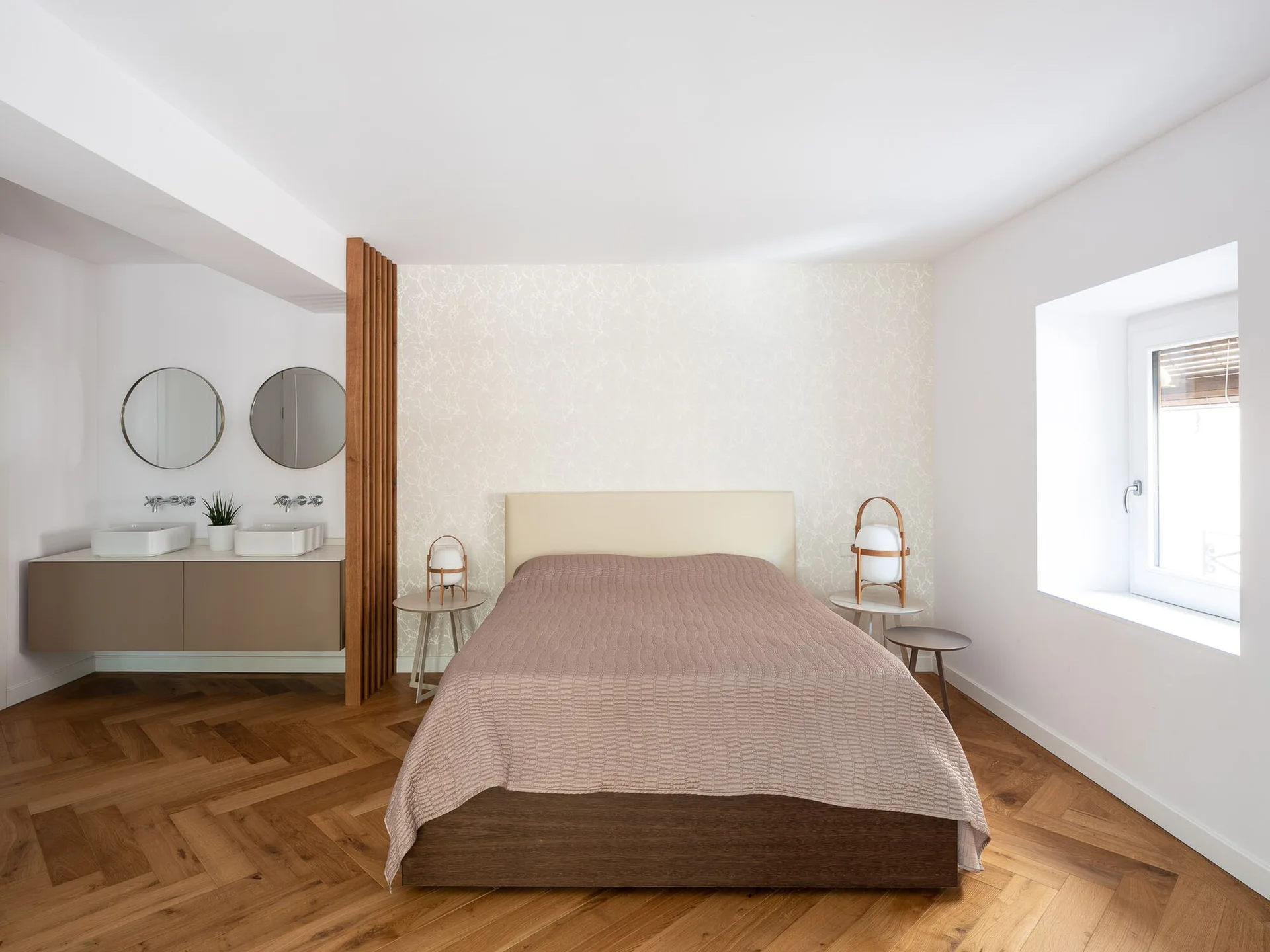 A minimalist bedroom featuring a bed with a light beige headboard, herringbone wood floors, and a bathroom vanity separated by a wood slat screen.