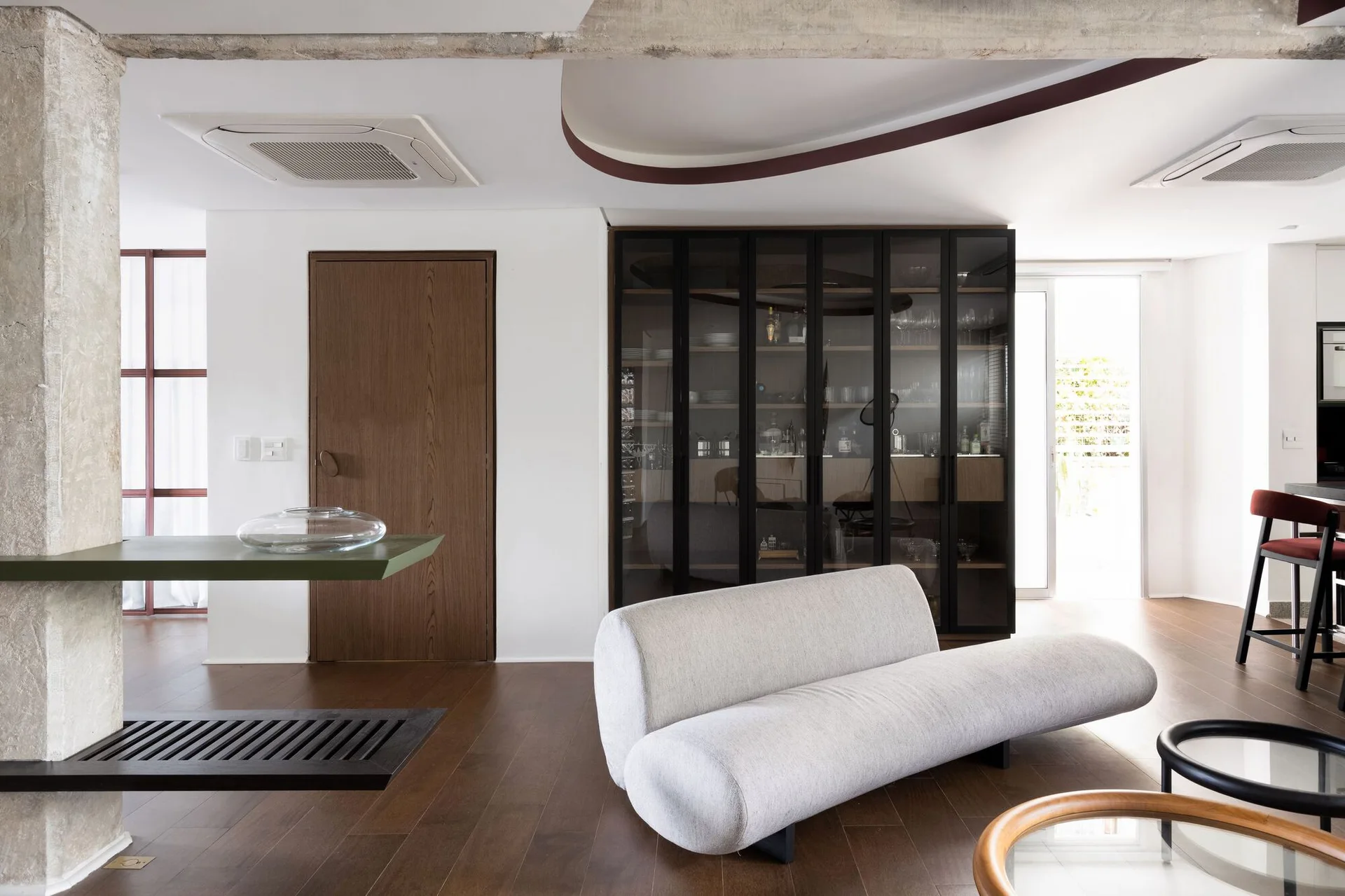 A modern living room features a curved grey sofa, dark wood flooring, and a tall glass-front display cabinet against a white wall.