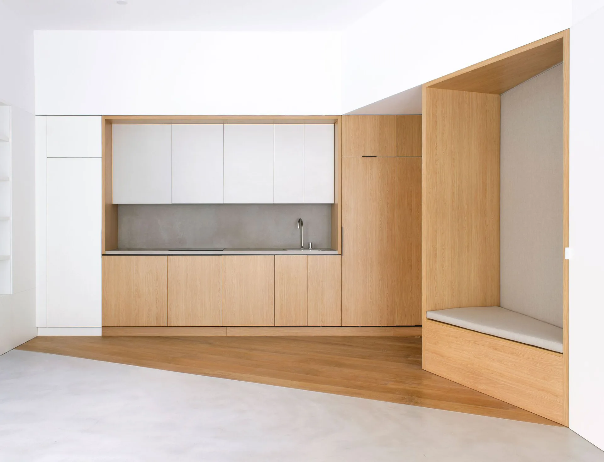 A minimalist kitchen features light wood and white cabinetry next to a recessed wooden seating nook on a diagonally transitioning floor.