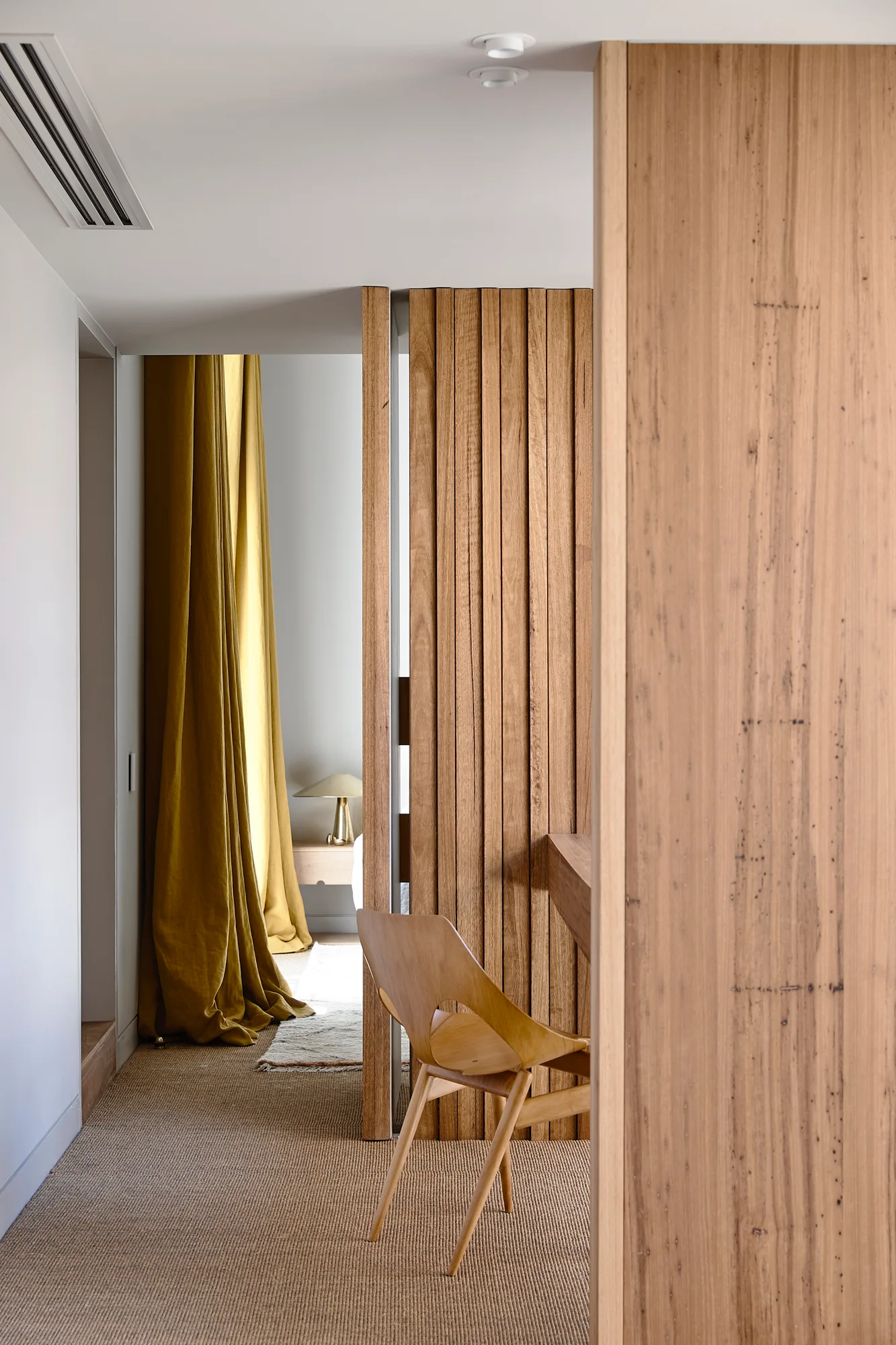 A view through a slatted wood partition wall shows a wooden chair, woven fiber flooring, and long yellow curtains in a minimalist bedroom.