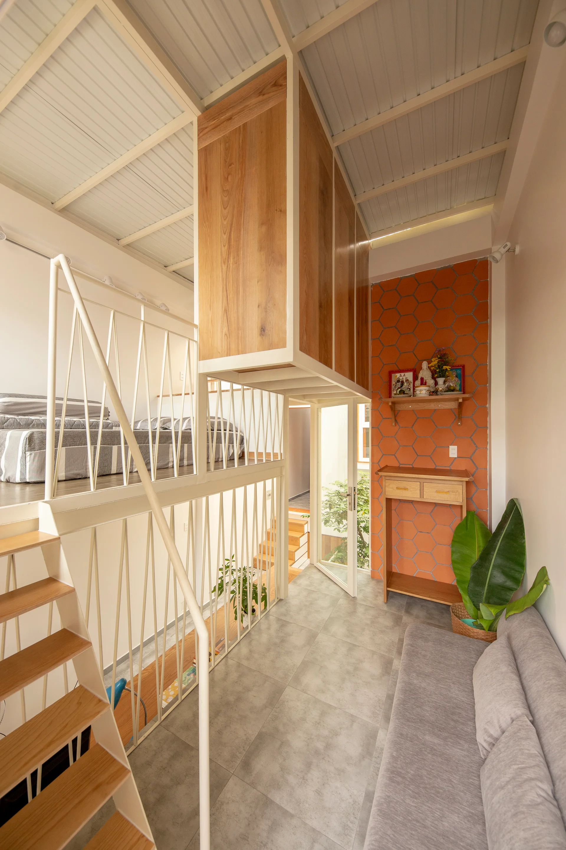 A compact loft apartment features a grey sofa, a white metal staircase leading to a loft bed, and a vibrant orange hexagonal tile accent wall.