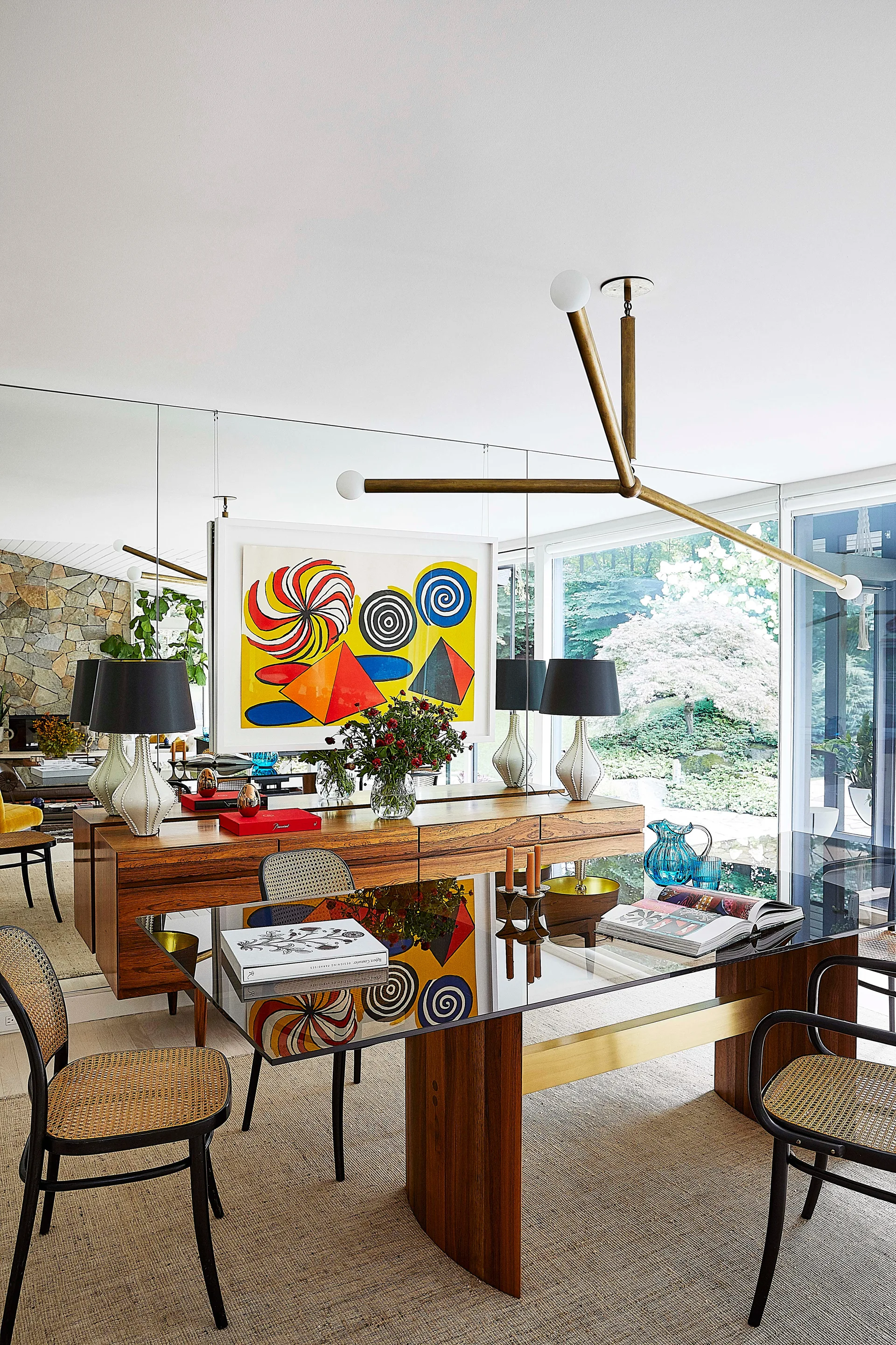 A dining room features a glass-topped table, cane-backed chairs, a wooden sideboard, and a full-height mirror wall reflecting colorful artwork.