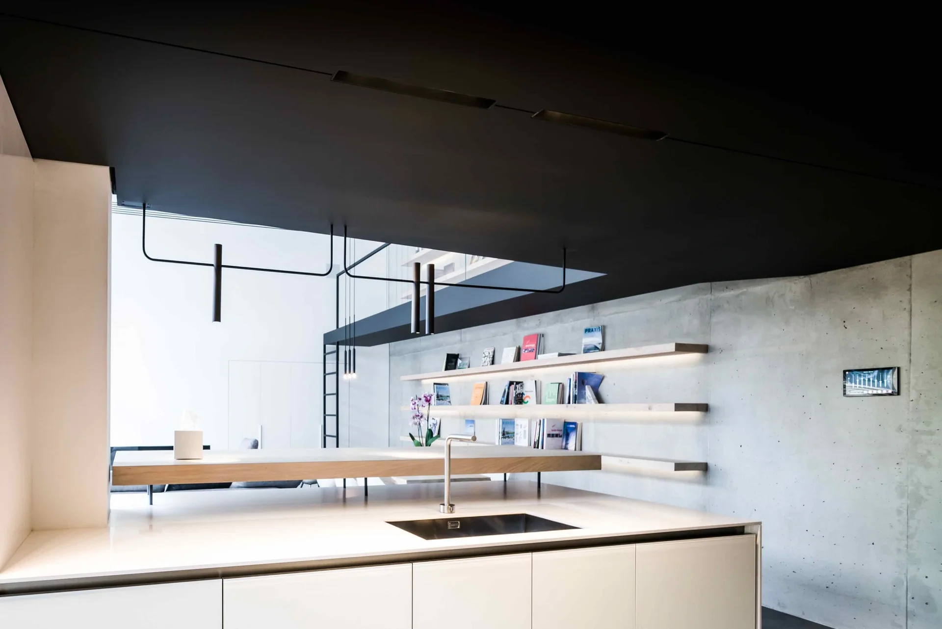 A modern kitchen featuring a white island with a sink set against an exposed concrete wall with three illuminated floating wooden shelves holding books.