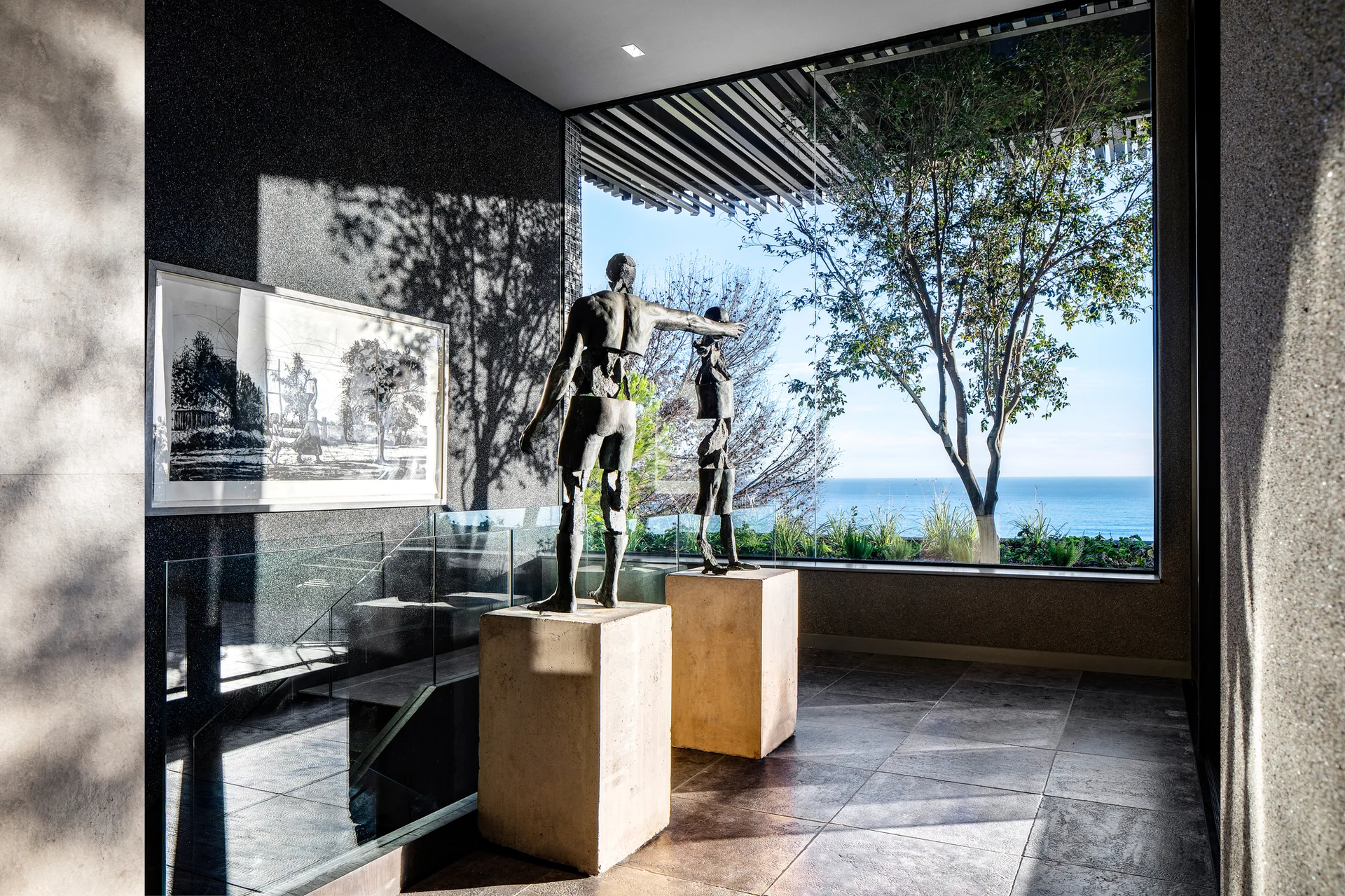 Two bronze figure sculptures stand on stone pedestals in a modern foyer next to a glass staircase and a large window overlooking the ocean.