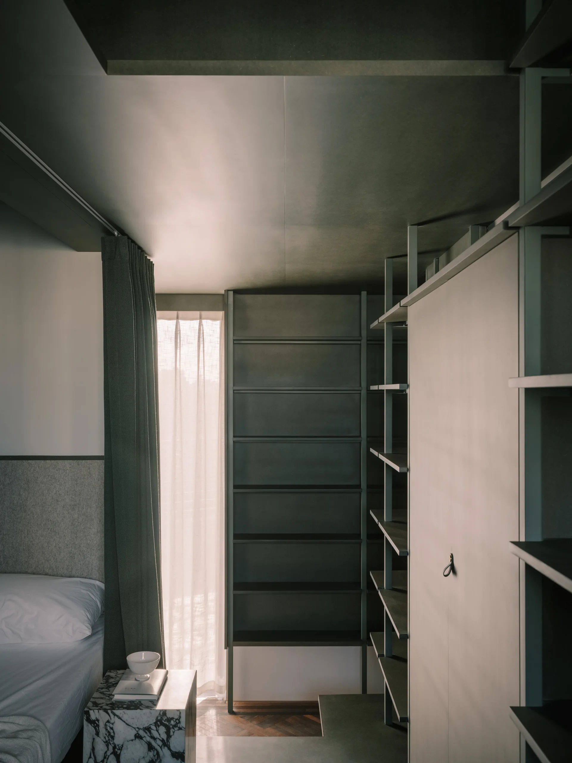 A modern bedroom features a dark metal modular closet system next to a window with sheer curtains and a black-and-white marble block side table.
