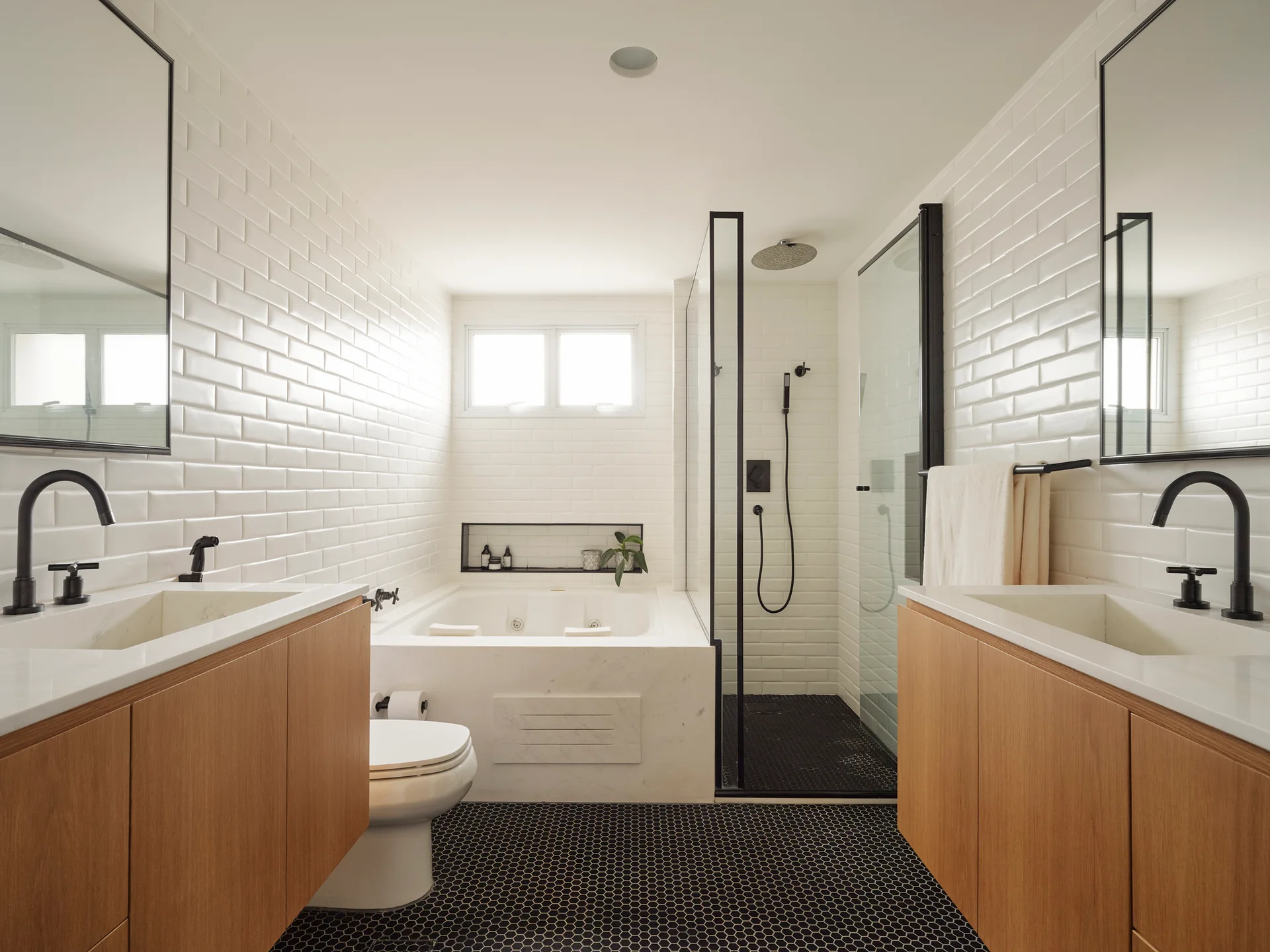 A symmetrical modern bathroom with opposing wood vanities, glossy white subway tiles, a built-in bathtub, and small black hexagonal floor tiles.