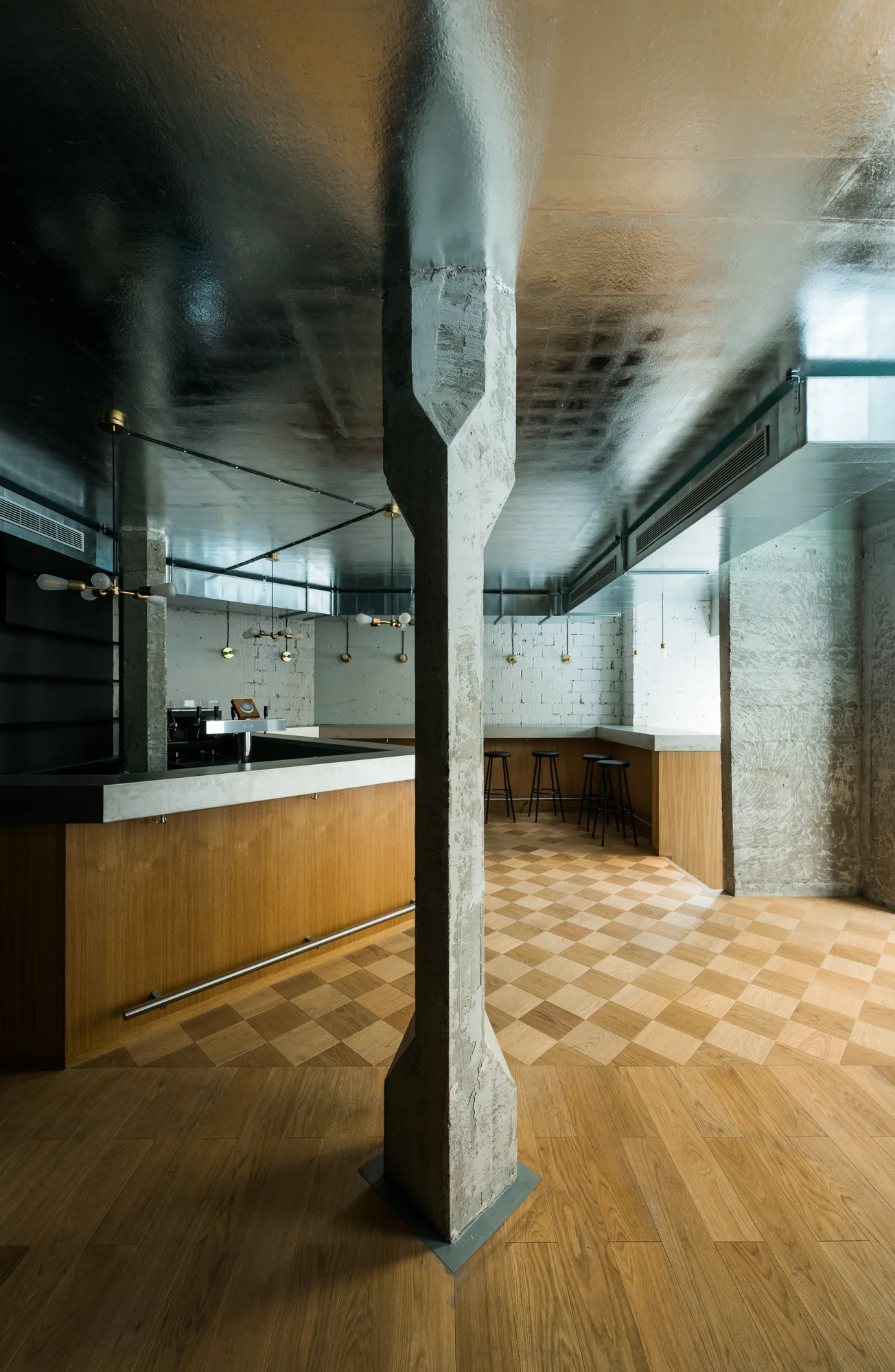 An industrial bar interior featuring a raw concrete pillar, diamond pattern parquet wood flooring, and a highly reflective silver metallic ceiling.
