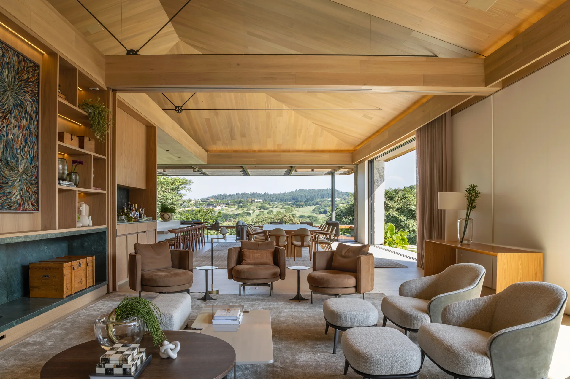A spacious living room featuring a sloped wood-plank ceiling, neutral armchairs, a built-in wooden bookcase, and an open wall leading to an outdoor terrace.