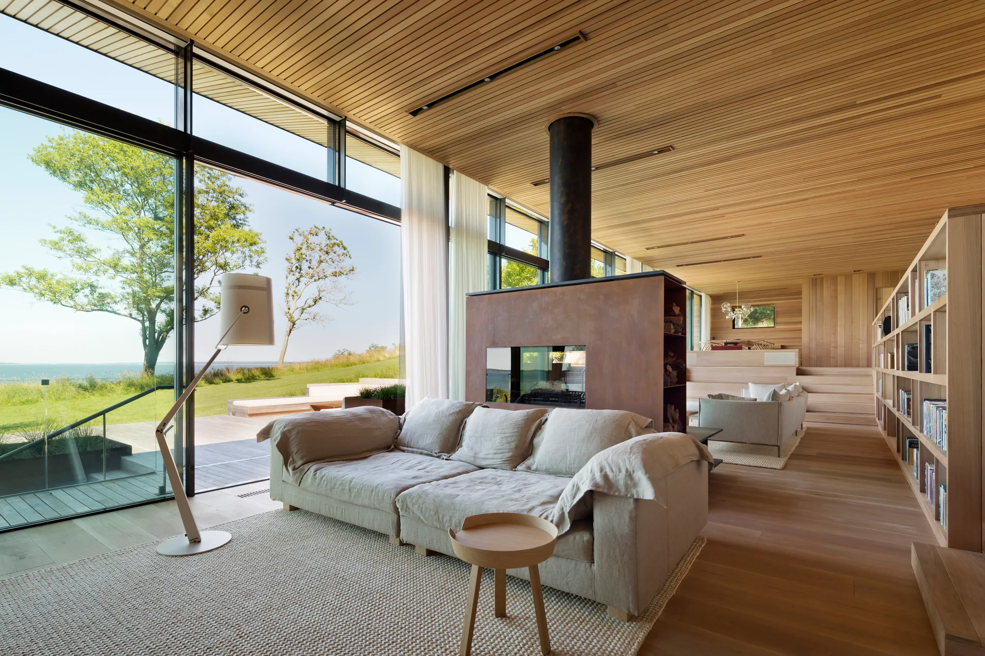 A modern living room featuring a slatted wood ceiling, a light linen sofa, a central fireplace, and large windows overlooking a waterfront.