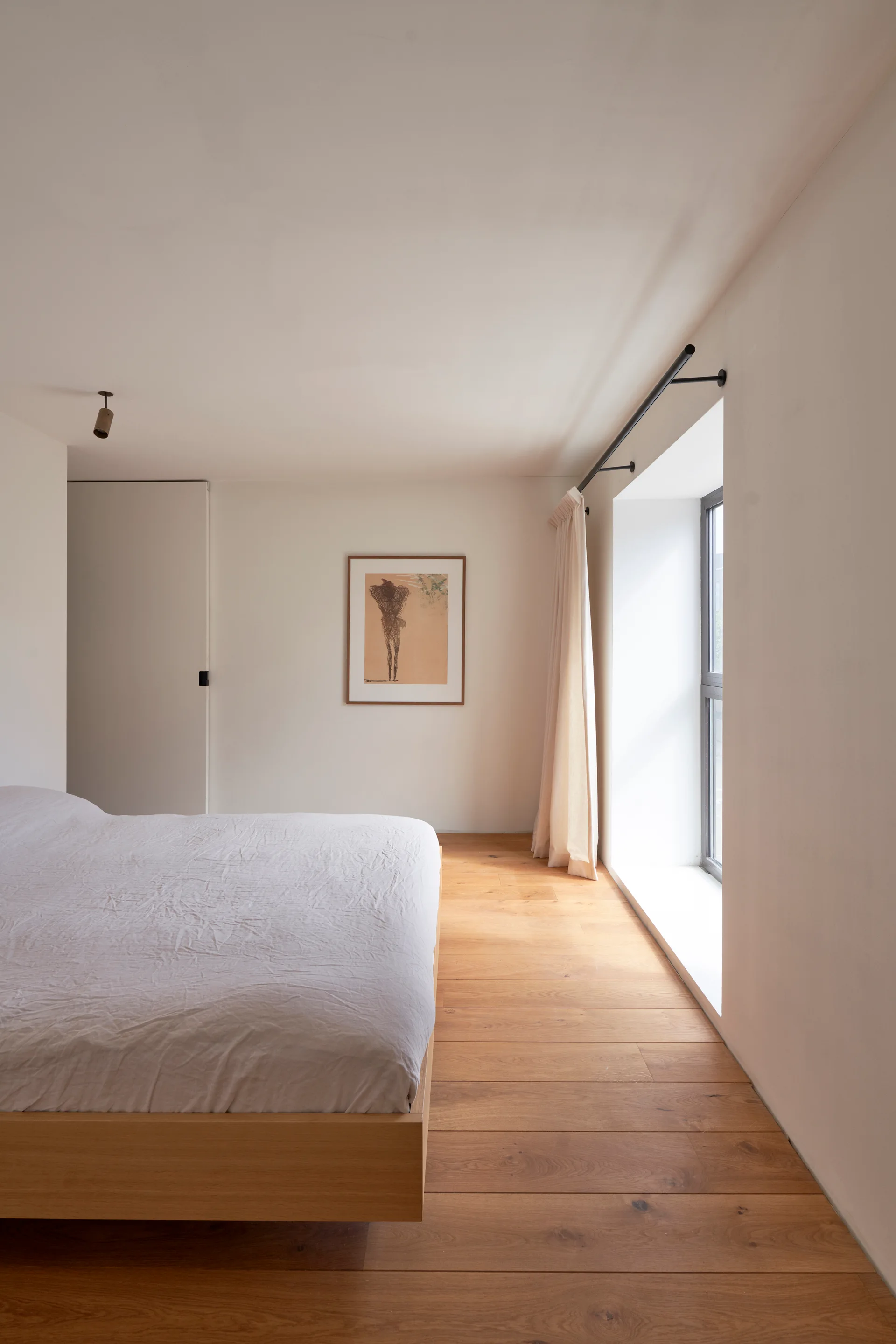 A minimalist bedroom featuring a low wooden platform bed, wide light oak floorboards, and a deeply recessed window with a single linen curtain.