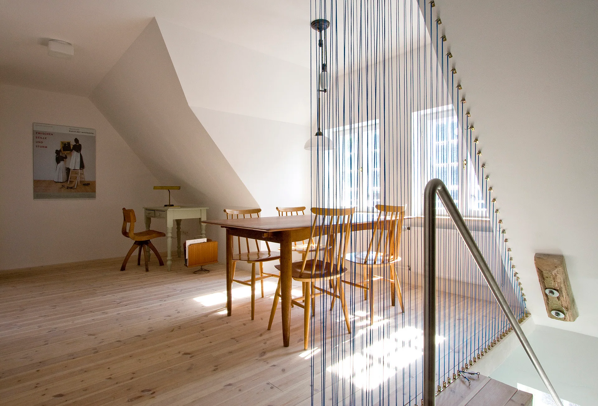 A bright attic dining room featuring a wooden table, chairs, and a staircase divider made of vertical blue cords against sloping white walls.