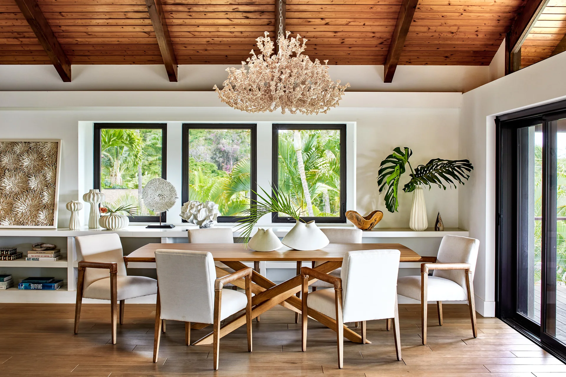 A dining room features a wooden table and upholstered chairs beneath a vaulted wood ceiling and a textured coral-style chandelier.