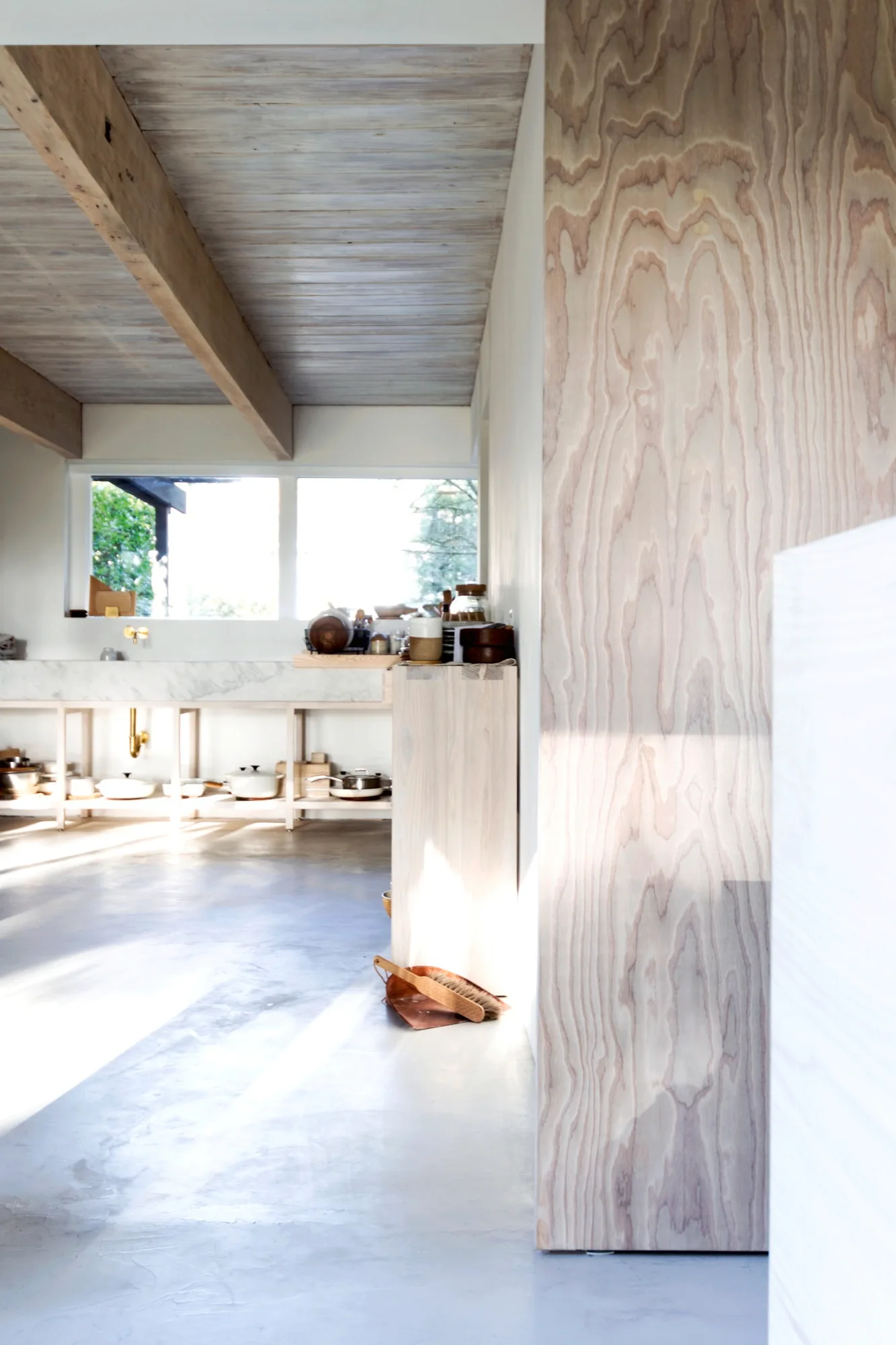 A bright minimalist kitchen features exposed wooden ceiling beams, a marble countertop over open shelves, and a plywood column in the foreground.