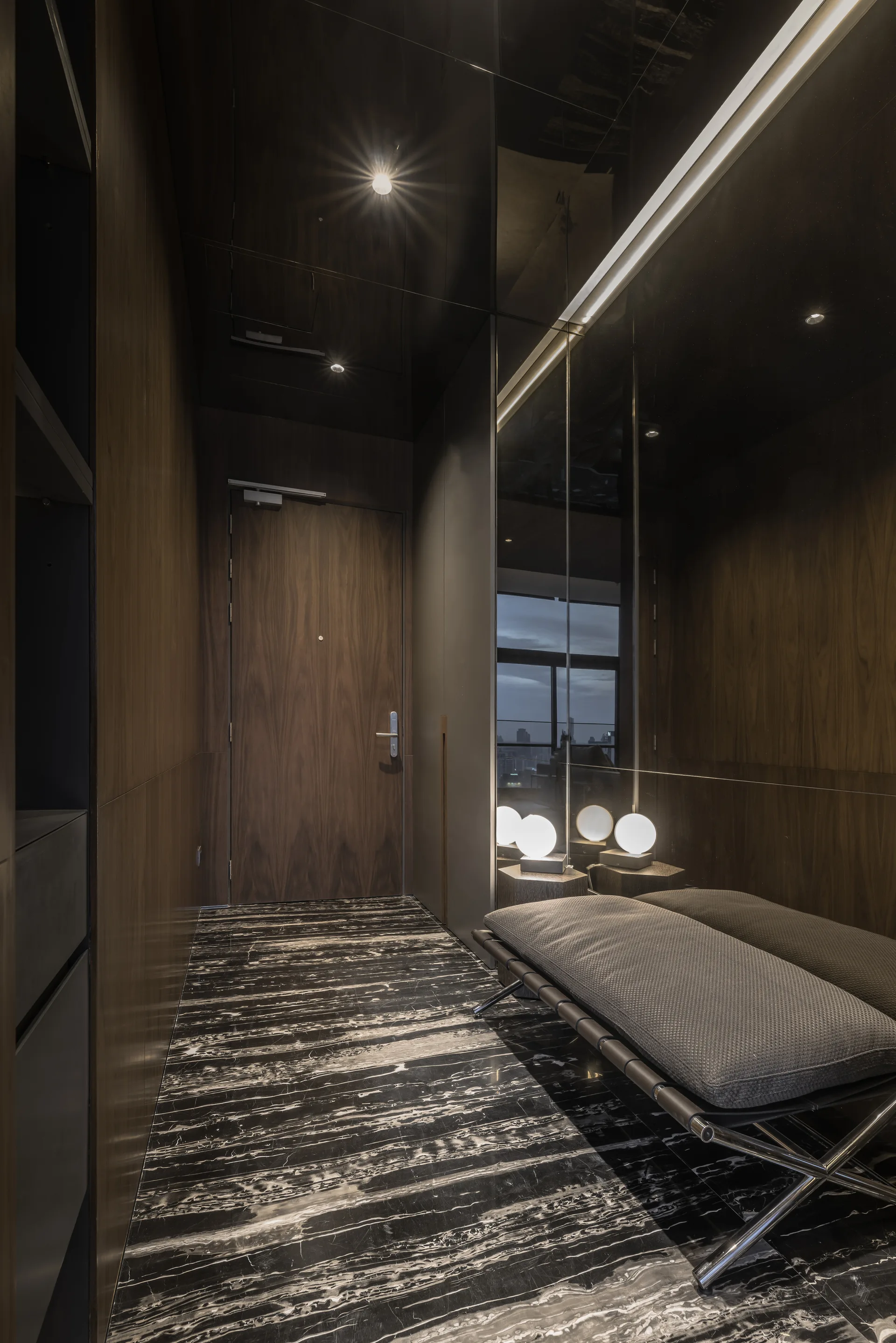 A modern residential entryway features dark wood-paneled walls, highly veined black marble flooring, a grey bench, and a large mirrored wall.