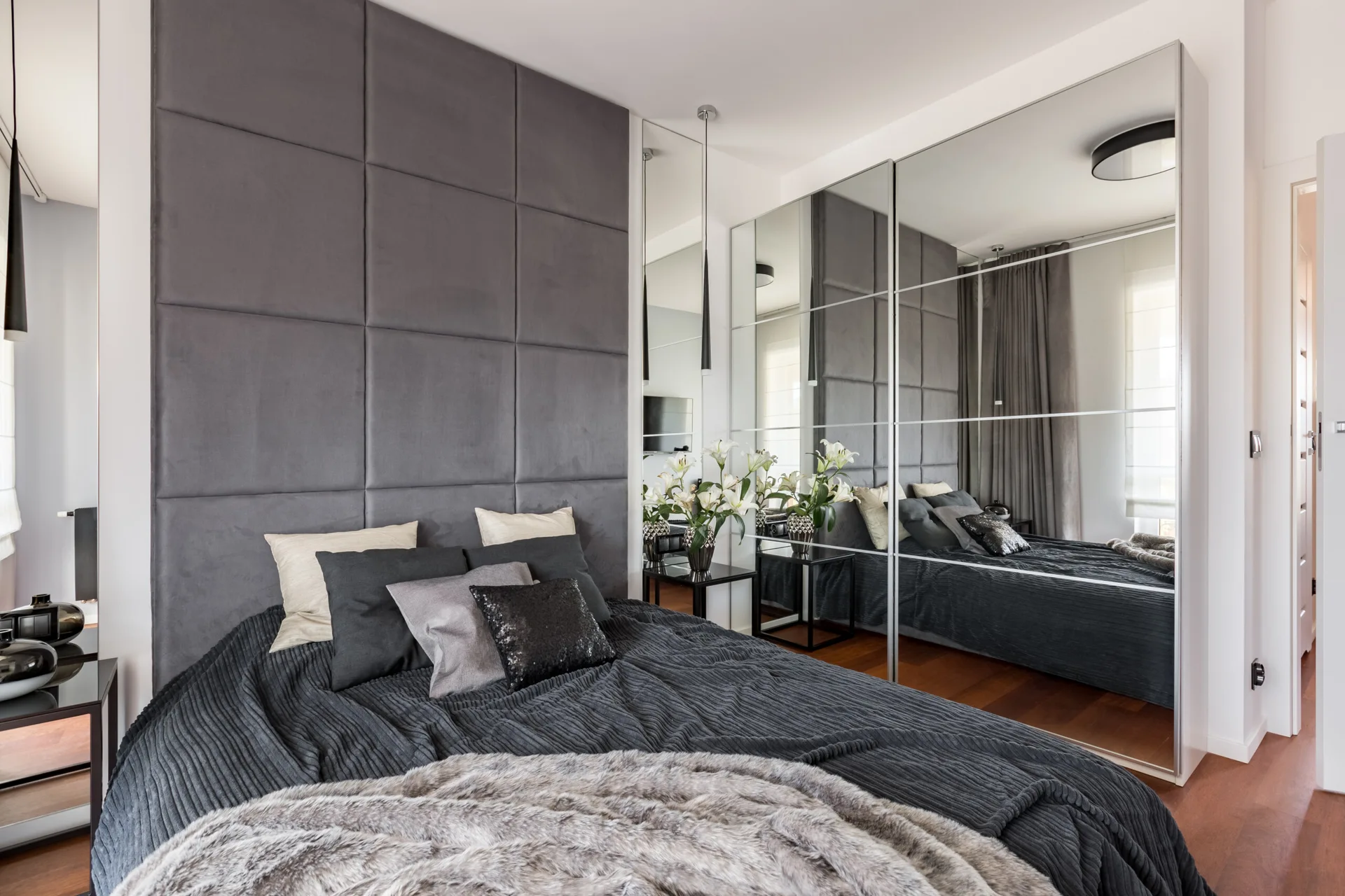 A modern bedroom features a grey upholstered panel headboard wall, textured dark bedding, and a large sliding mirrored closet reflecting the room.