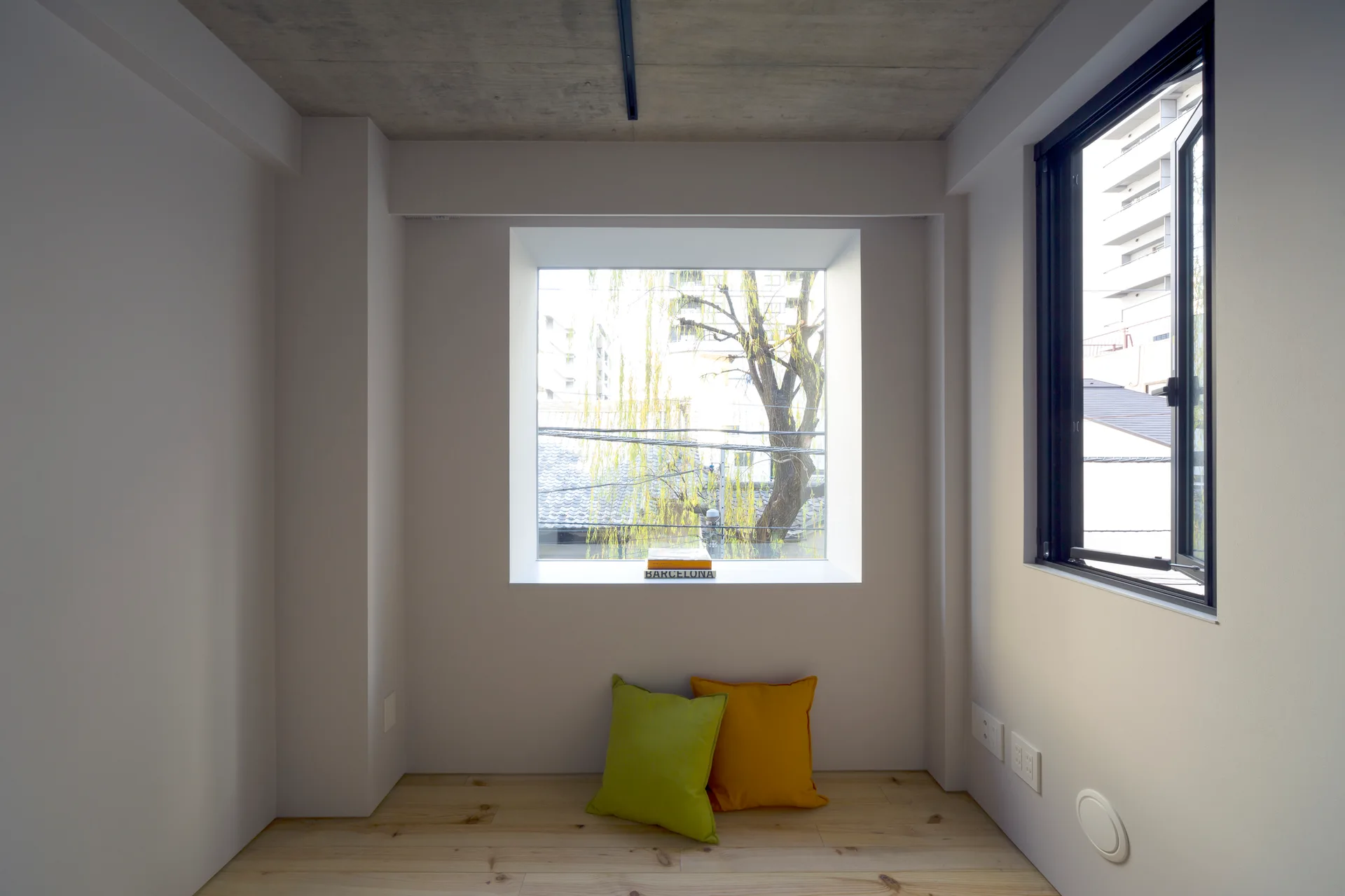 A minimalist room with an exposed concrete ceiling, light wood floors, a large square window, and two colorful floor cushions.