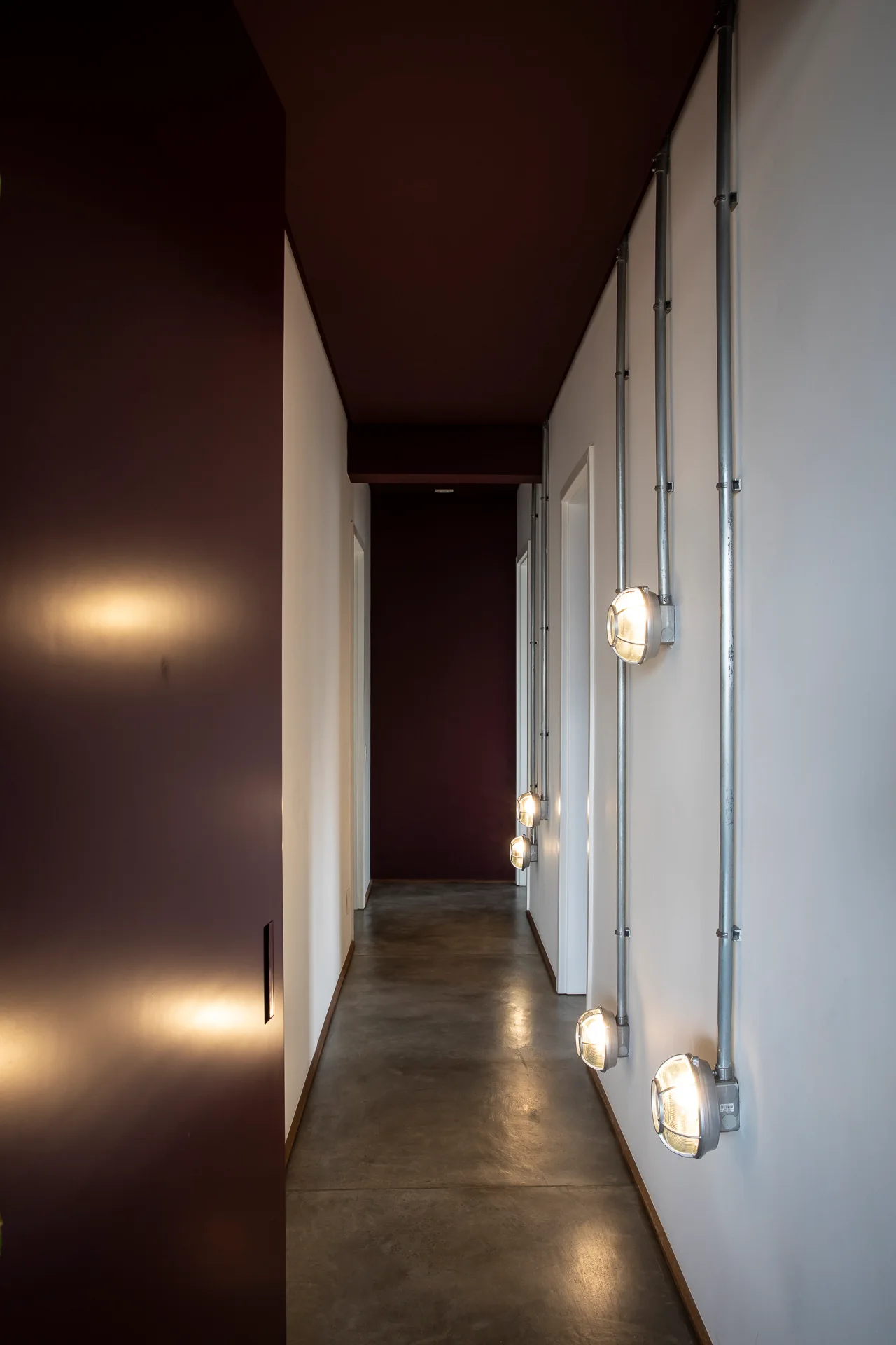 A long narrow hallway features polished concrete flooring, white and dark red walls, and industrial wall sconces with exposed metal conduits.