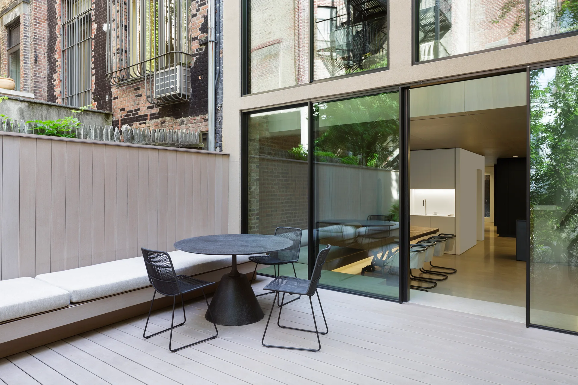 A modern urban patio features a circular black table, wire mesh chairs, and a long built-in bench next to sliding glass doors opening to a kitchen.