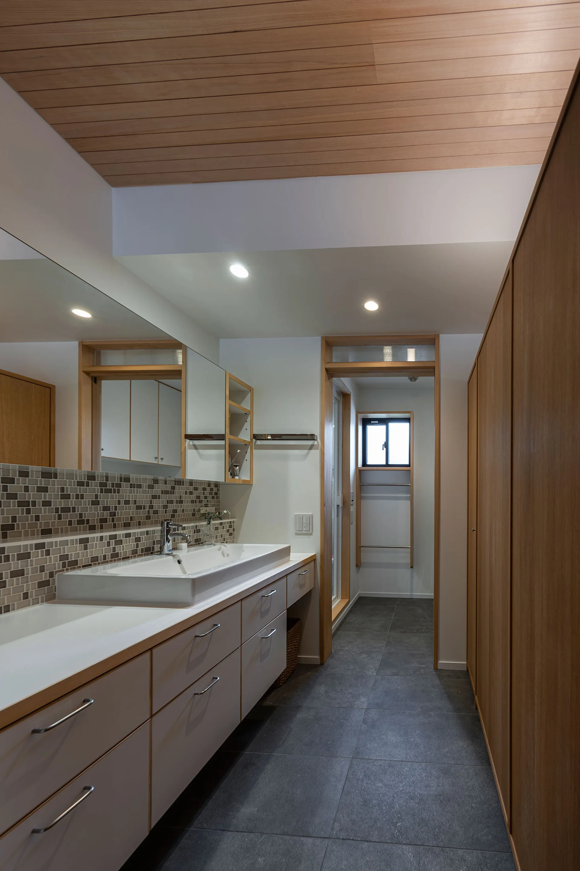 A modern bathroom features a long white vanity with a mosaic tile backsplash, dark grey floor tiles, and a natural wood plank ceiling.