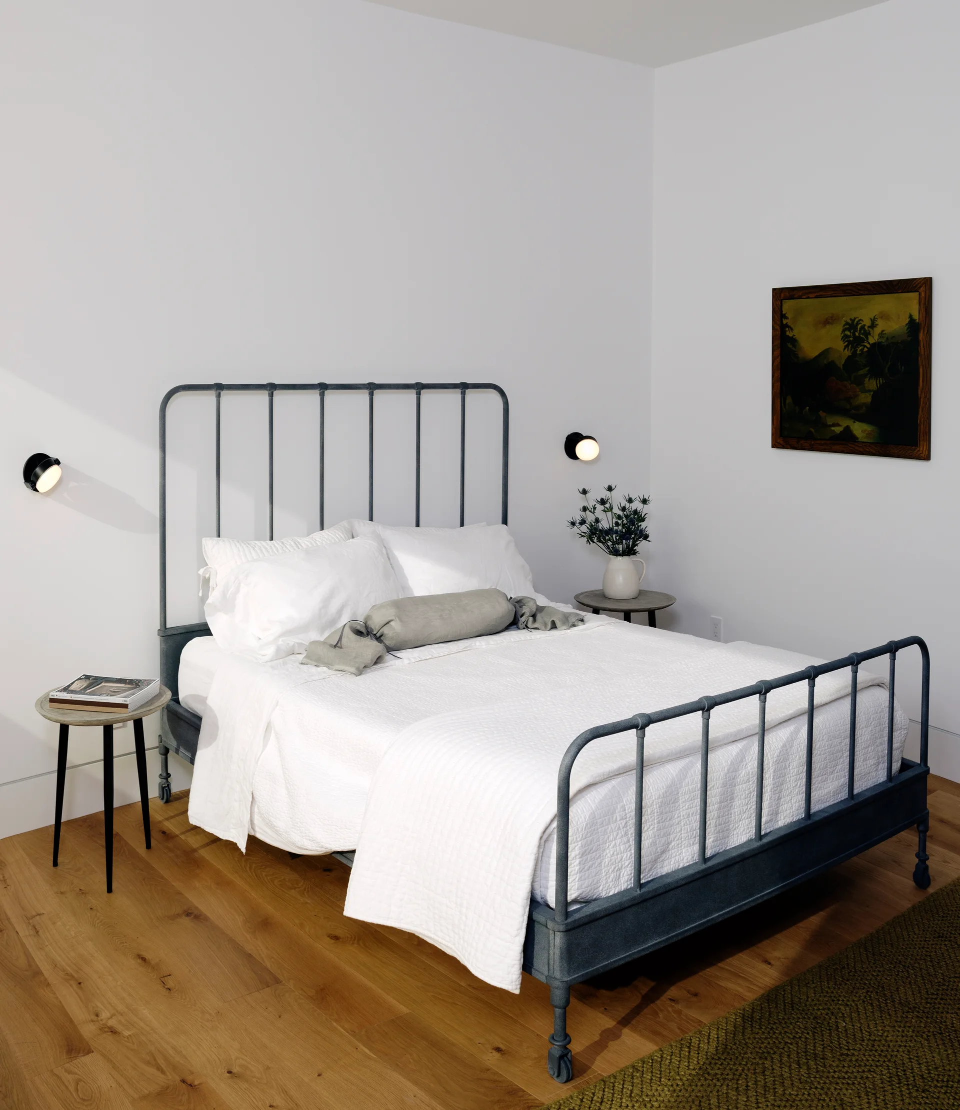 A minimalist bedroom featuring a charcoal metal bed frame with white bedding, small round wooden nightstands, and spherical wall sconces.