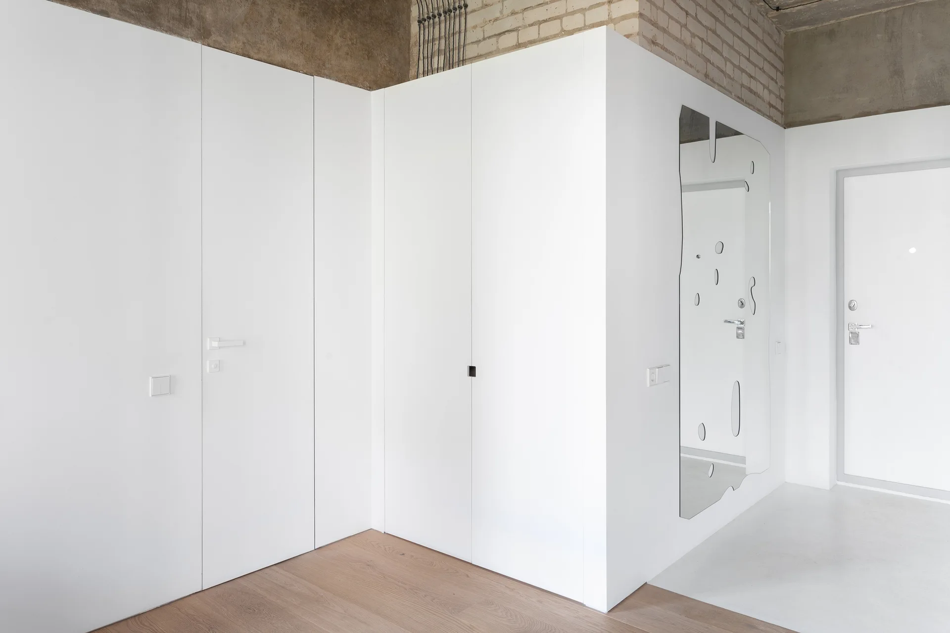 A minimalist entryway features smooth white floor-to-ceiling built-in storage closets, light wood flooring, and an exposed raw concrete ceiling.