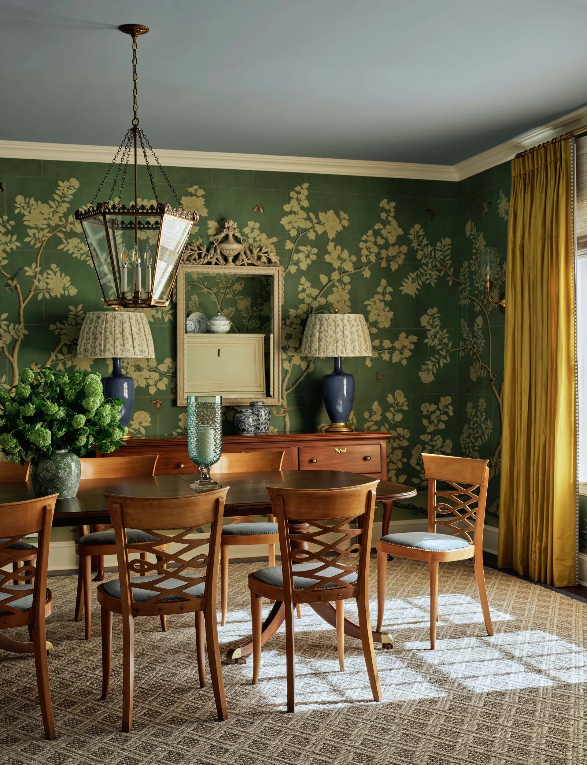 A traditional dining room featuring wooden lattice-back chairs, a dark wood table, green botanical wallpaper, and a blue painted ceiling.