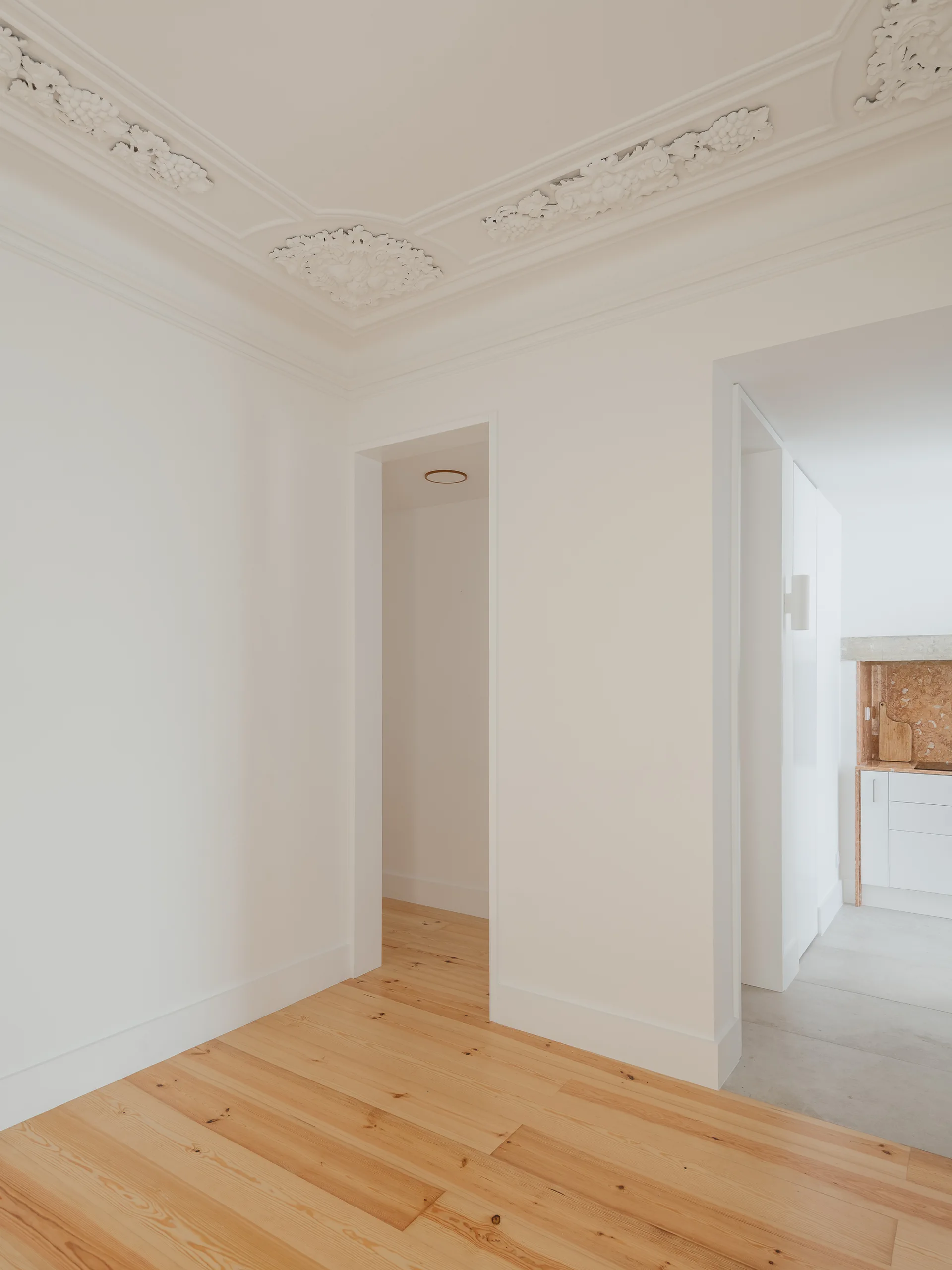 A bright hallway features light wood floors, smooth white walls, and a highly detailed ornate white plaster ceiling with decorative reliefs.