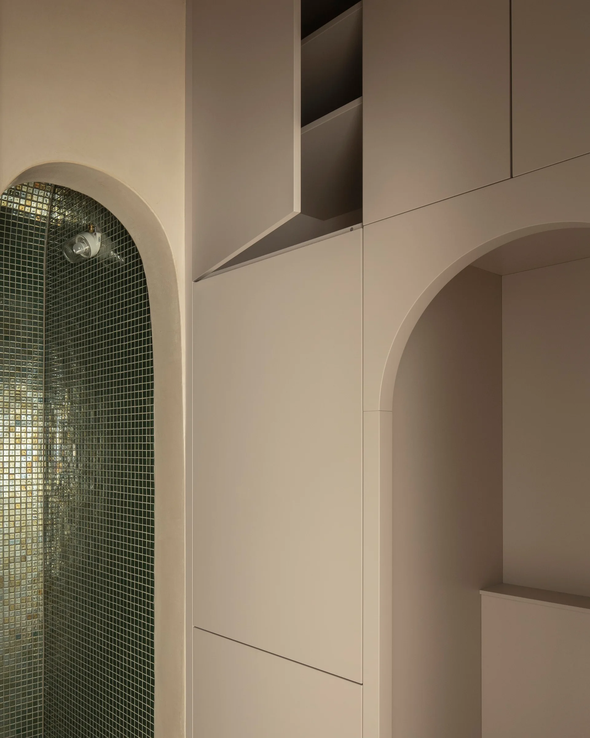 A bathroom featuring a tiled arched shower alcove with green mosaic tiles next to custom beige floor-to-ceiling cabinets with an integrated arch.