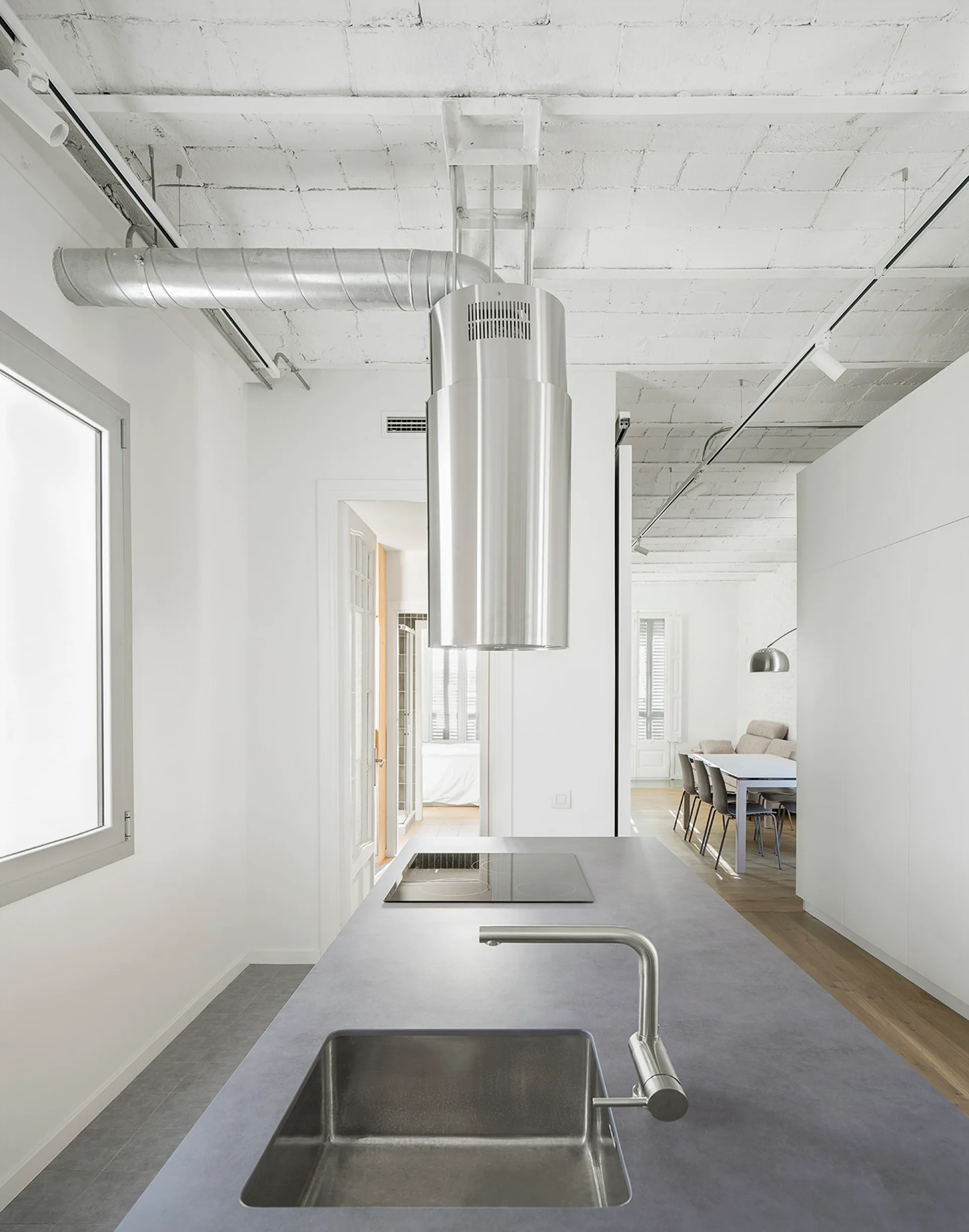 A minimalist loft kitchen features a gray concrete-look island with a sink under a cylindrical stainless steel range hood and a white vaulted brick ceiling.