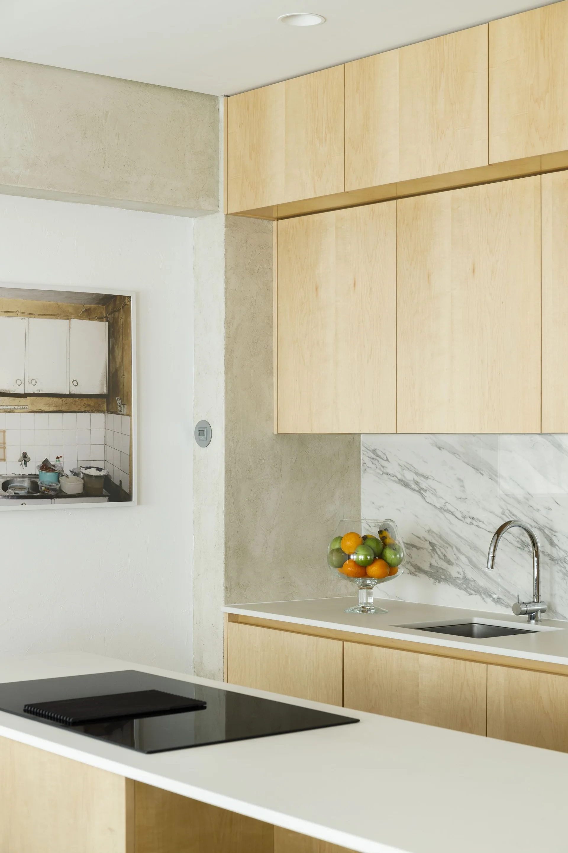 A minimalist kitchen features light wood cabinets, a veined marble backsplash, a stainless steel faucet, and a concrete-textured pillar.
