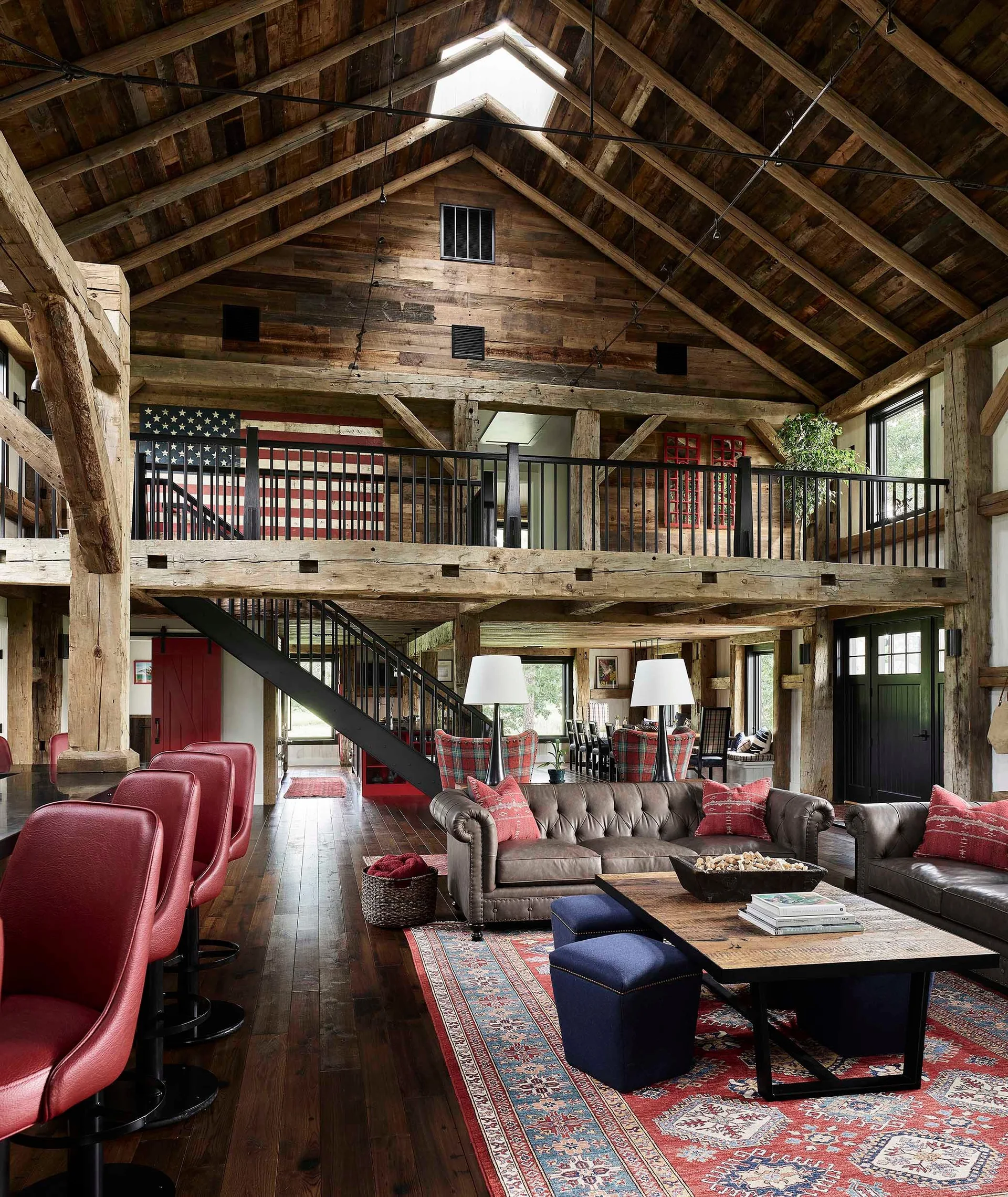 A timber-frame barn conversion living room features tufted leather sofas, red bar stools, a wooden coffee table, and a lofted mezzanine.