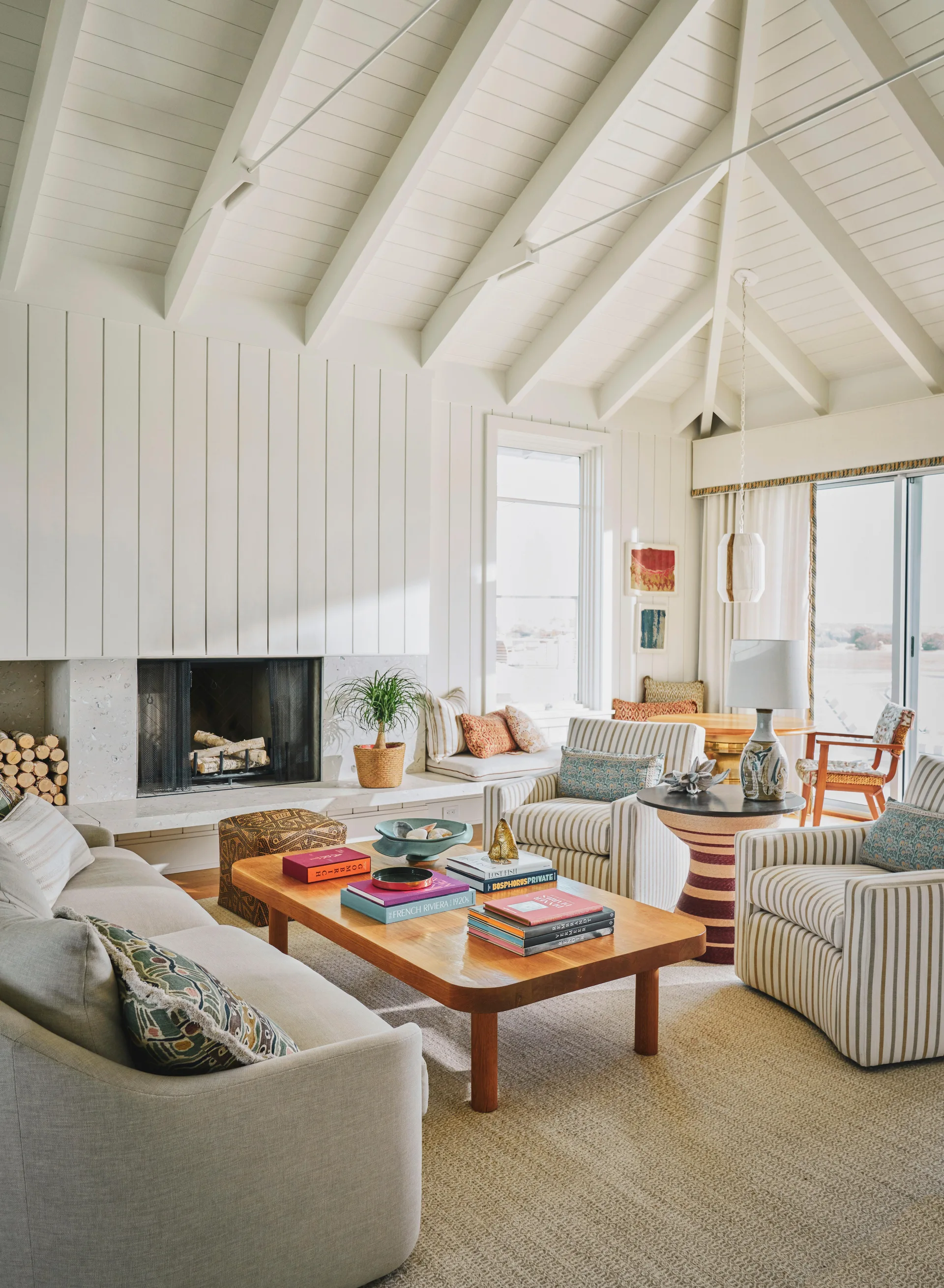 A bright living room features a vaulted beam ceiling, a stone fireplace, striped armchairs, and a wooden coffee table with books.