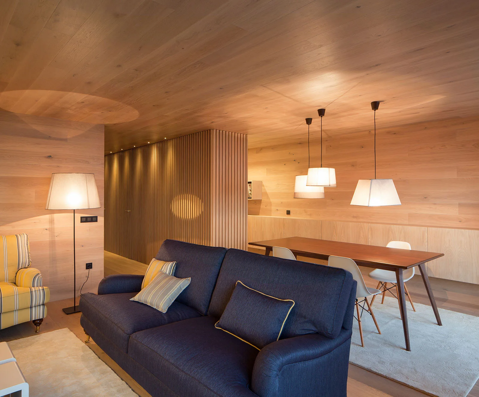 A modern living space features wood-paneled walls and ceiling, a navy blue sofa, and a wooden dining table with white chairs.