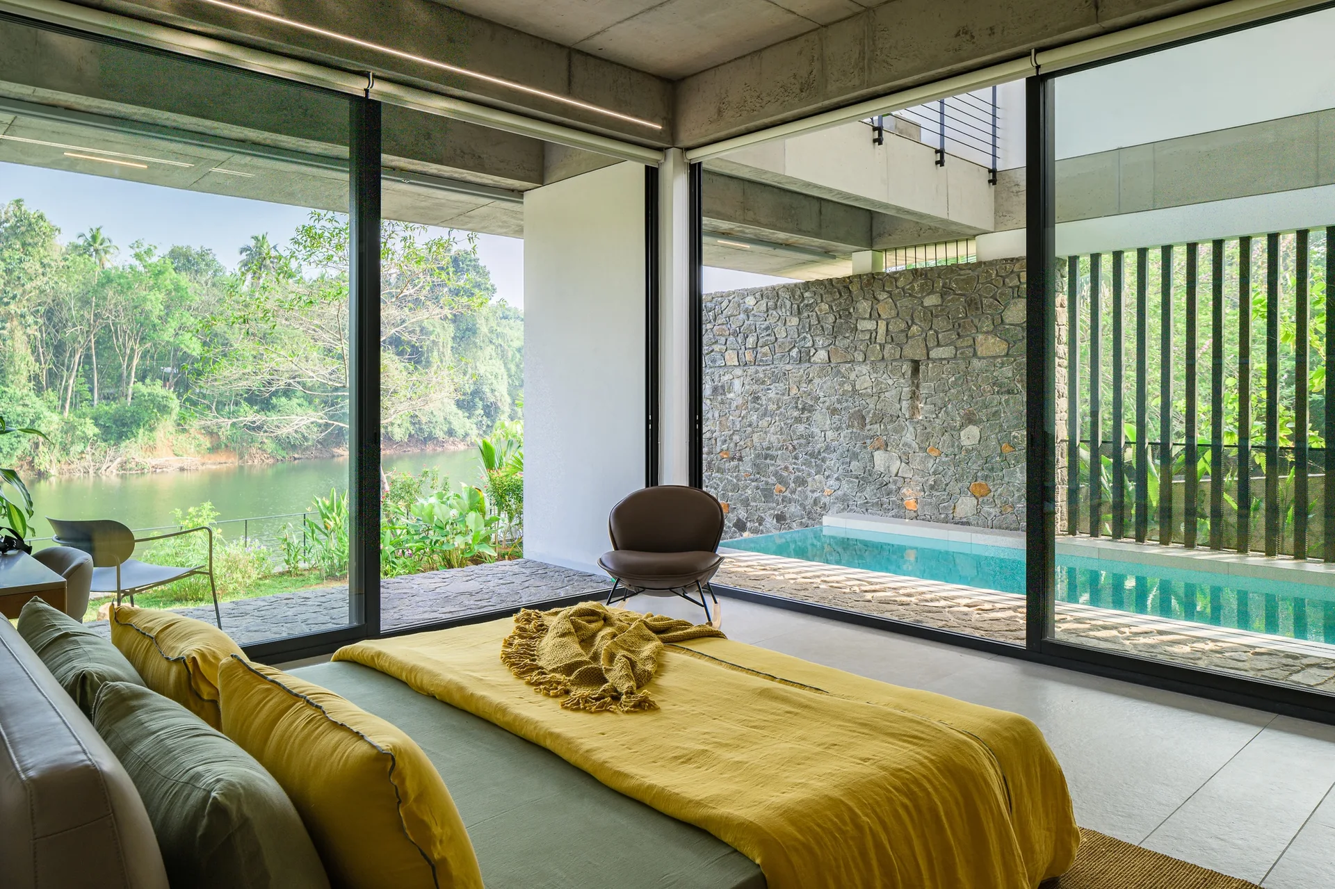 A modern bedroom features an ochre yellow bed and sliding glass doors that open to a private outdoor pool and a stone wall.
