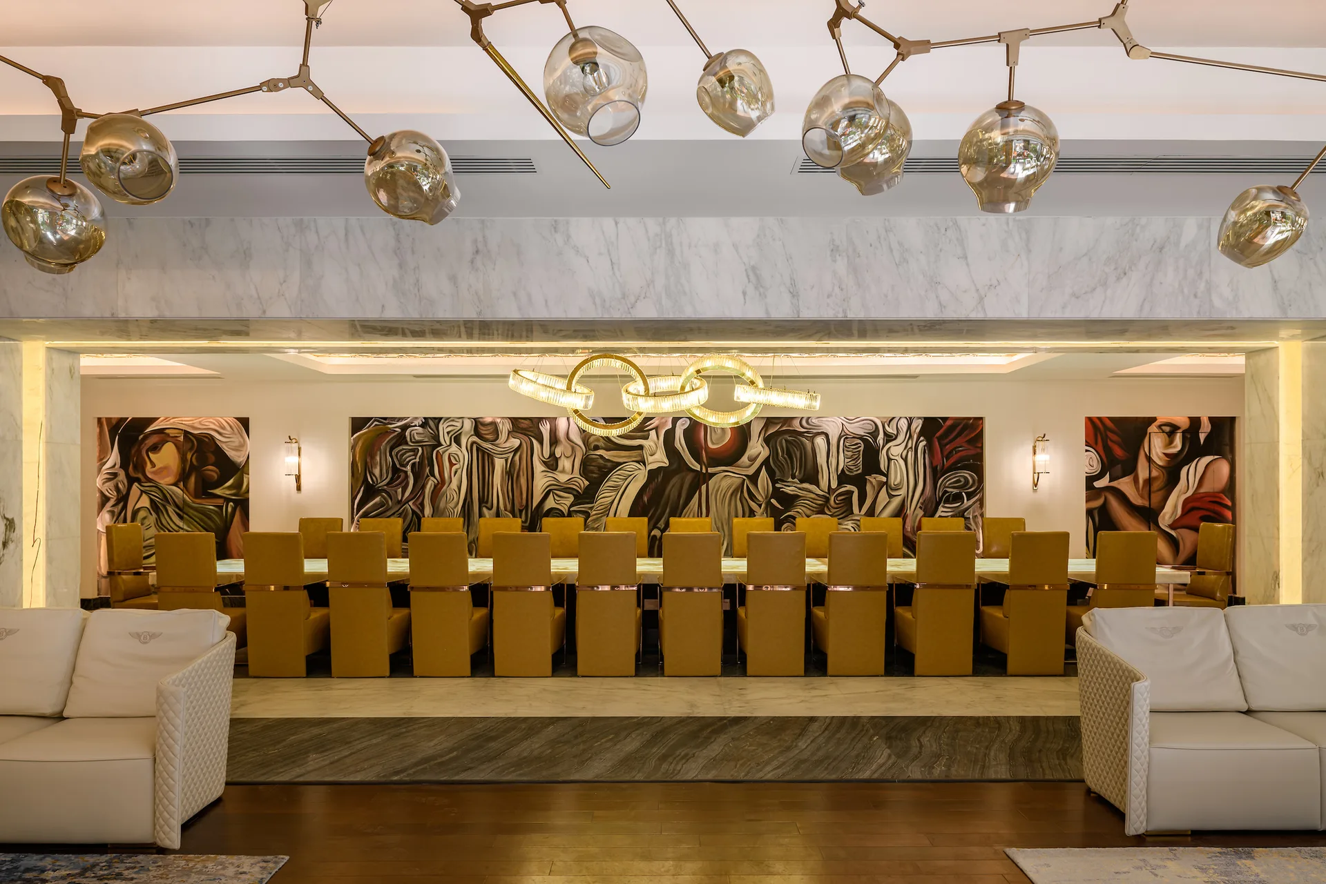 A long illuminated dining table surrounded by mustard yellow chairs sits below a branching glass chandelier and large abstract wall paintings.