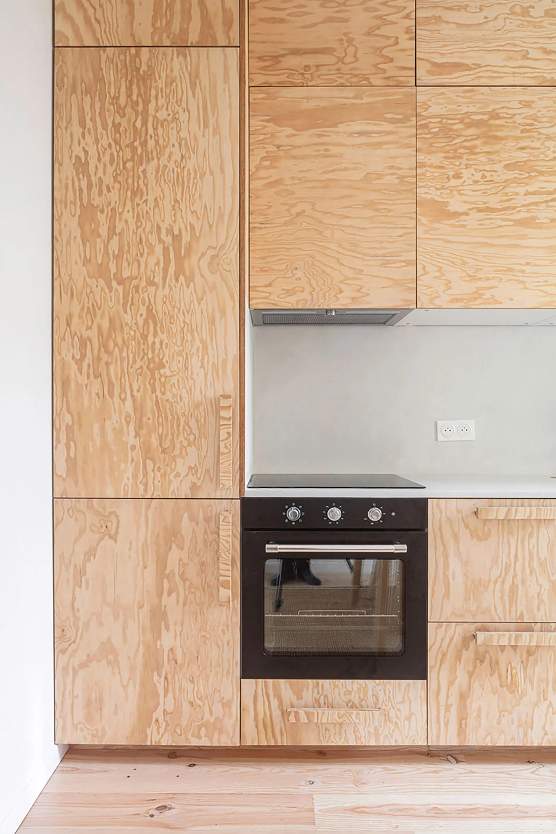 A close-up of light plywood kitchen cabinets with integrated handles and a black built-in oven.