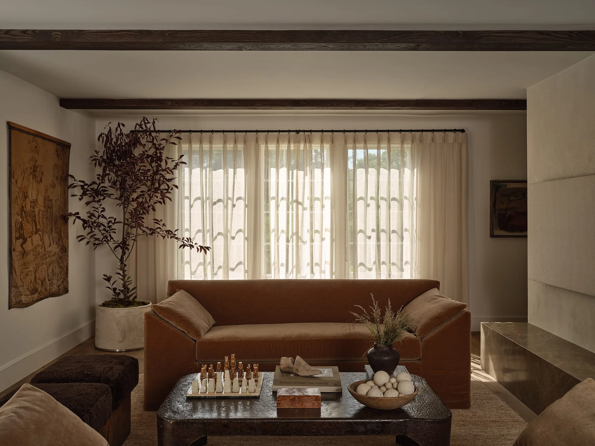 A warm living room featuring a brown velvet sofa, a textured dark wood coffee table, sheer cream curtains, and dark exposed ceiling beams.