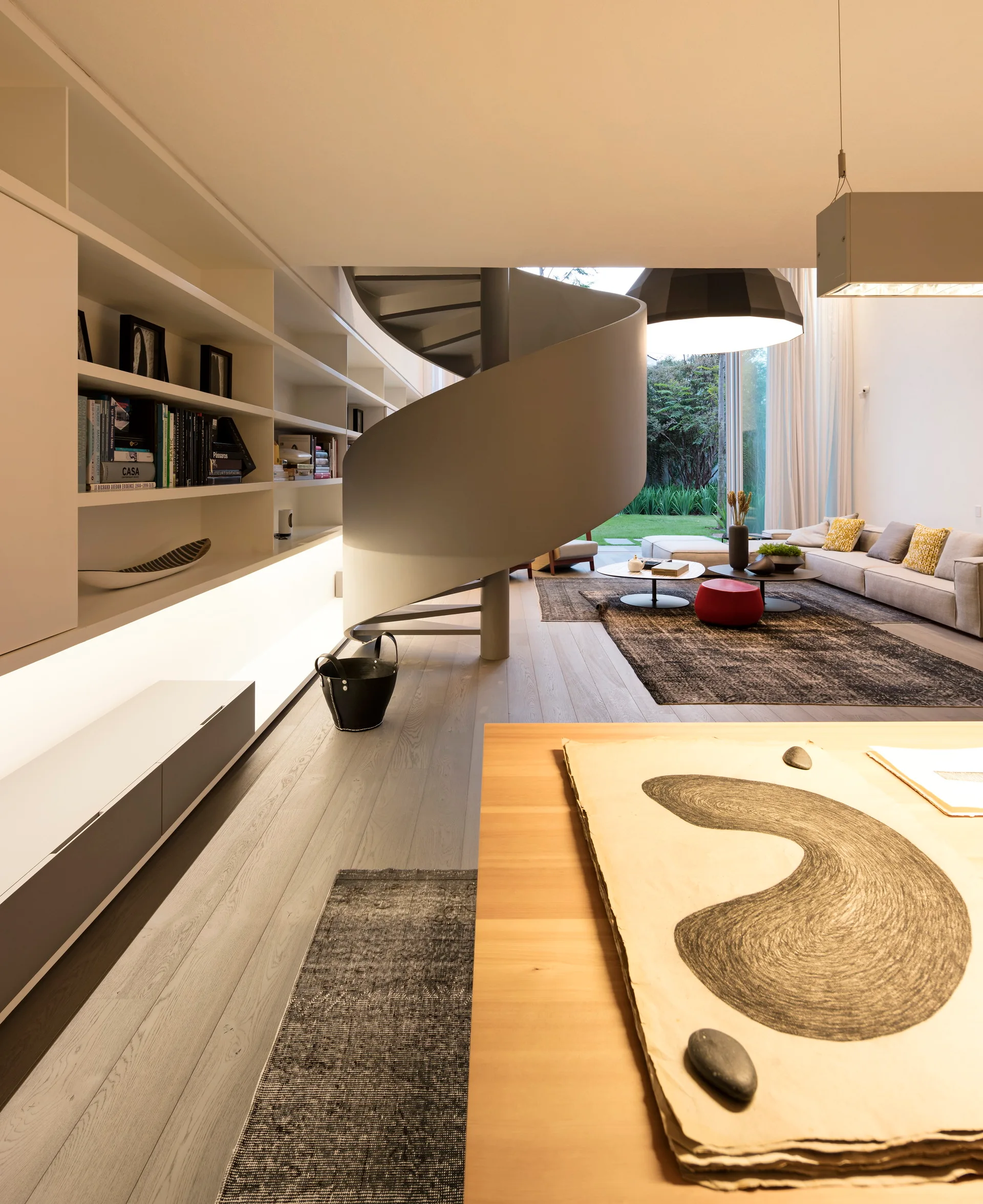 A modern living room features a sculptural grey spiral staircase, a long white built-in bookshelf with integrated lighting, and a beige sofa.