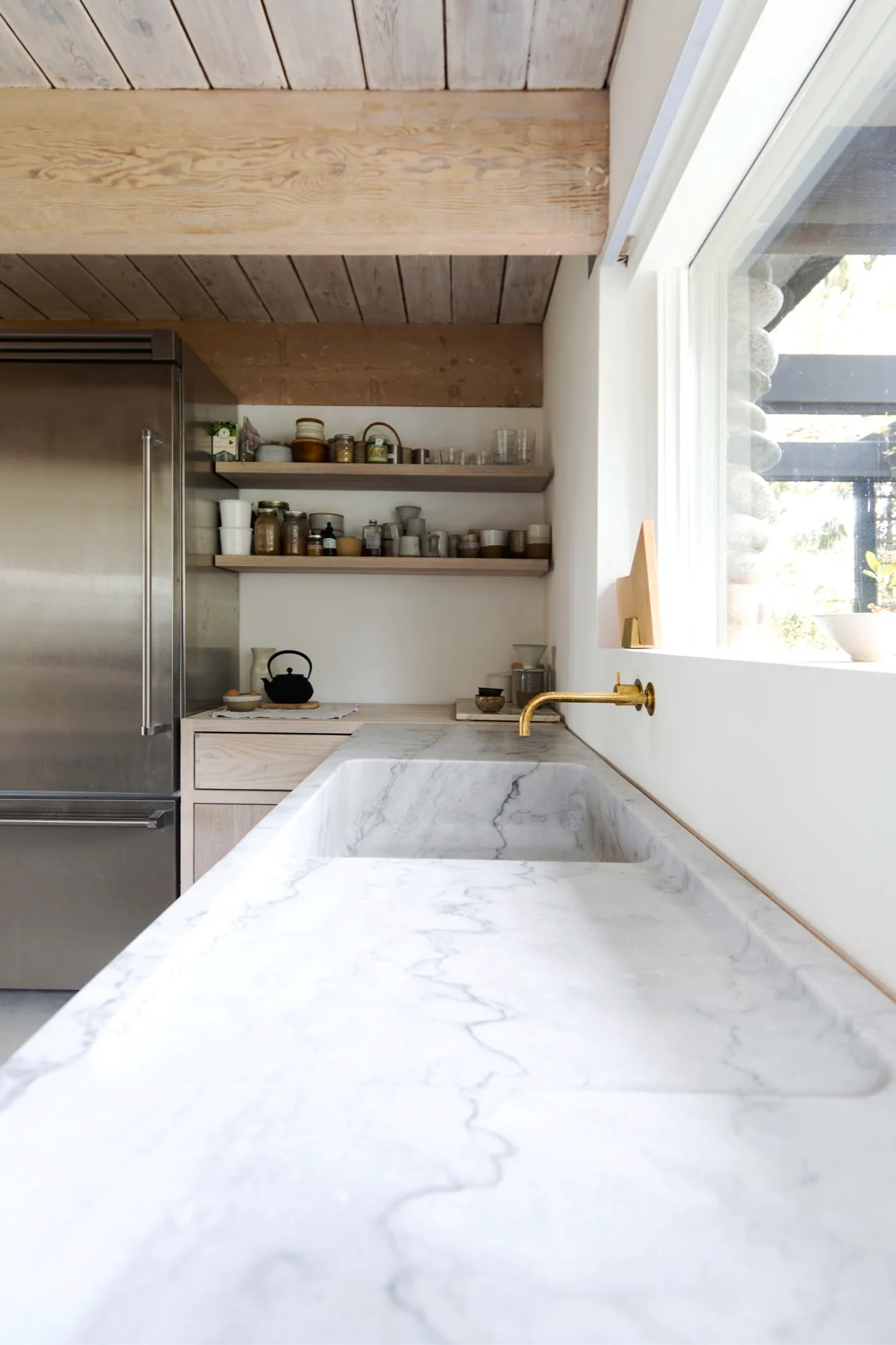 A minimalist kitchen featuring an integrated marble sink with a brass wall-mounted faucet beneath open wood shelving and a beamed wood ceiling.