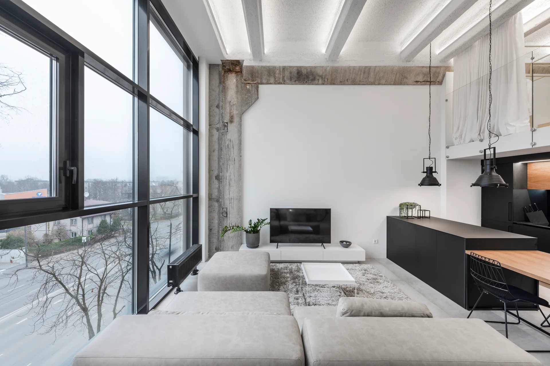 A loft living room with a light gray sectional sofa, an exposed concrete pillar, and a large grid window overlooking trees.