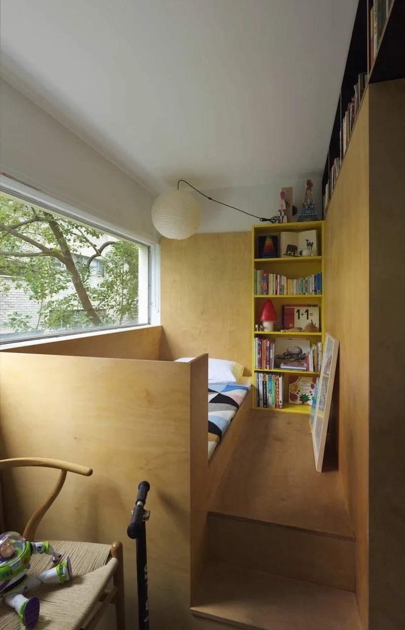 A small children's bedroom features a built-in plywood platform bed next to a window, a bright yellow bookcase, and a paper globe pendant light.