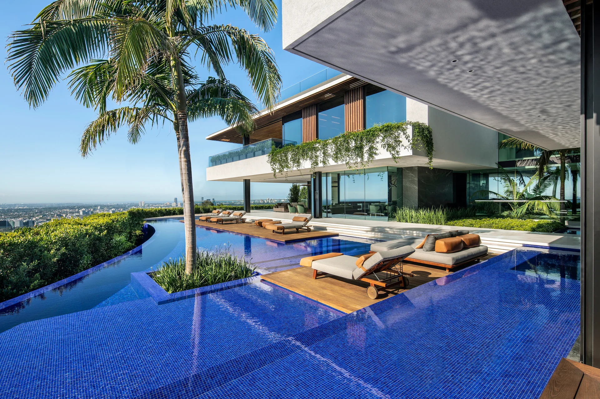 A modern white villa features a bright blue tiled infinity pool with wood lounge decks and a tall palm tree overlooking a city view.